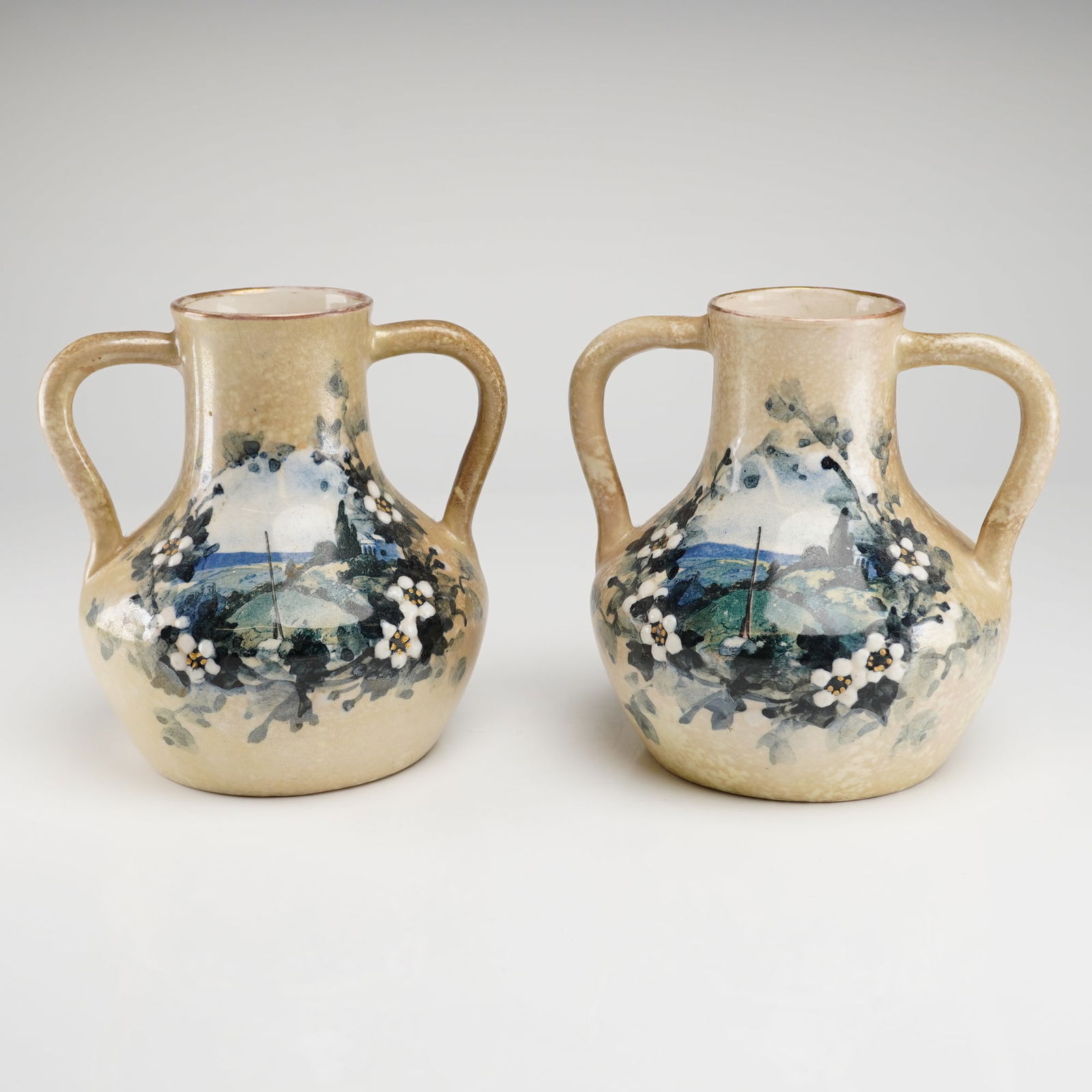 A Pair of Alfred Stellmacher Teplitz Floral Earthenware Vases: Alfred Stellmacher Teplitz earthenware vases. The vessels are amphora-form, featuring a bulbous body, short neck, and opposing loop handles. The body is covered in a mottled, speckled beige-tan glaze.