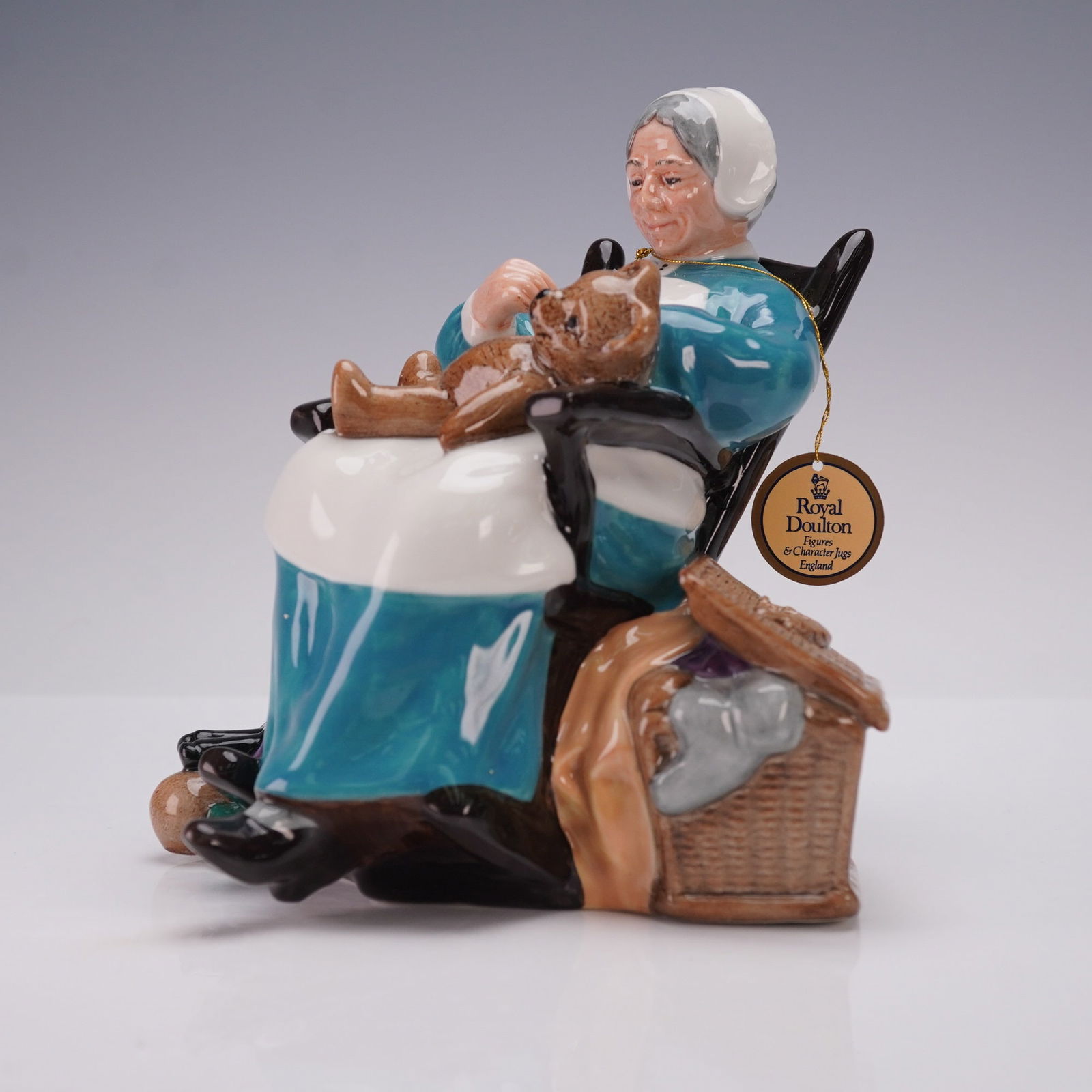 Royal Doulton Figurine, The Nanny HN2221: The hand painted porcelain piece depicts a seated nanny in a rocking chair holding a teddy bear on her lap, with toys including a drum, doll, and ball placed at her feet and a basket at her side. The