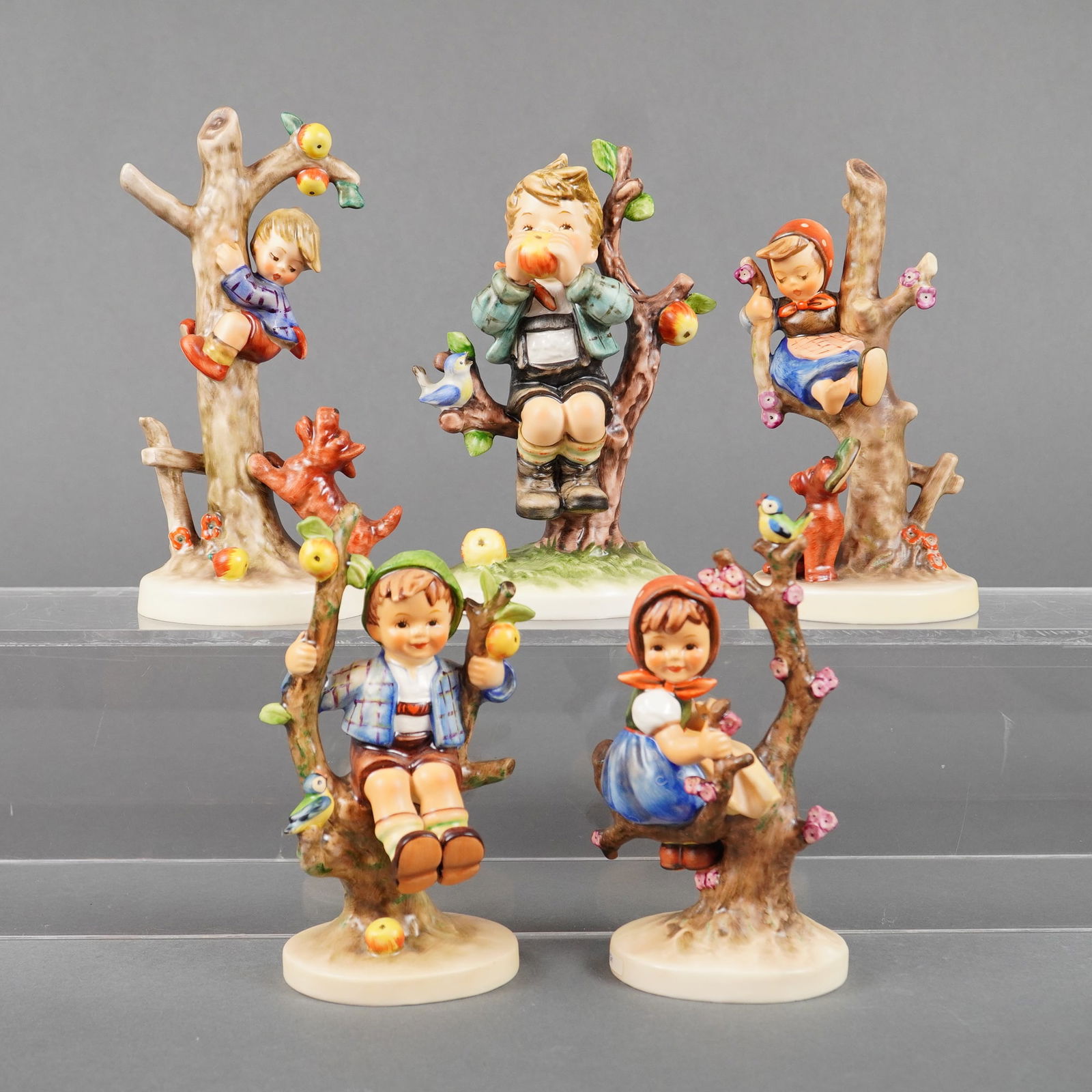 A Collection of 5 Goebel M.I. Hummel Tree Sitter Figurines: A collection of five glazed porcelain figural groups, identified as Goebel M.I. Hummel figurines from the 'Apple Tree' or 'Tree Sitter' series. Each piece depicts a young boy or girl seated among the