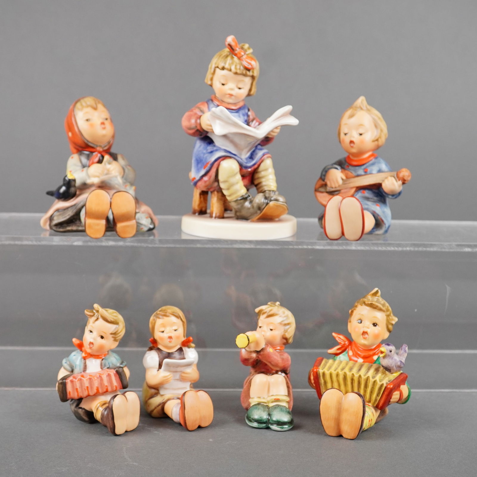 7 Goebel Hummel Figurines incl. What's New: A collection of 7pc Goebel Hummel figurines, executed in glazed ceramic. The collection depicts children engaged in various musical and recreational activities, including reading, singing, and playing
