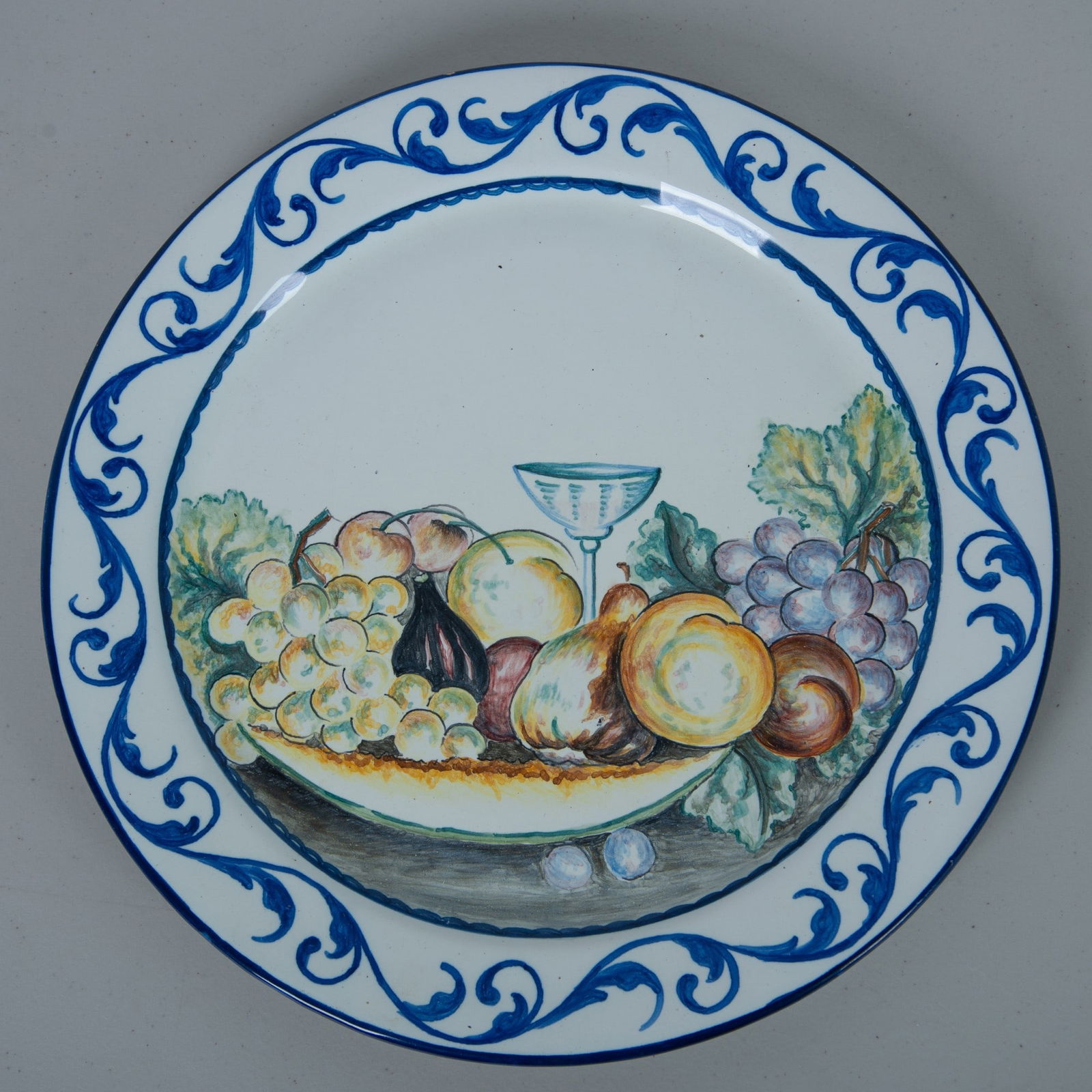Vintage Large Ceramic Fruit Wall Plate: A white hand painted decorative plate depicting an array of fruits, leaves and a wine glass, with a dark blue scrollwork pattern around the rim. It has two holes on the back to allow for wall hanging.