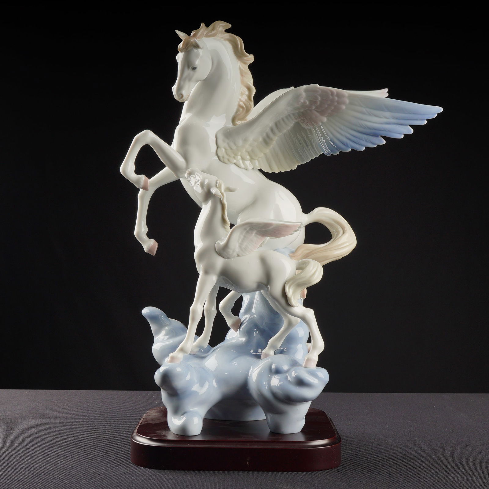 Lladro Porcelain Sculpture Pegasus 01001778 with Base: The glossy white porcelain depicts a rearing adult Pegasus and a smaller winged foal standing atop stylized pale blue clouds. The sculpture features soft accent coloration in pale blues, pinks, and ta