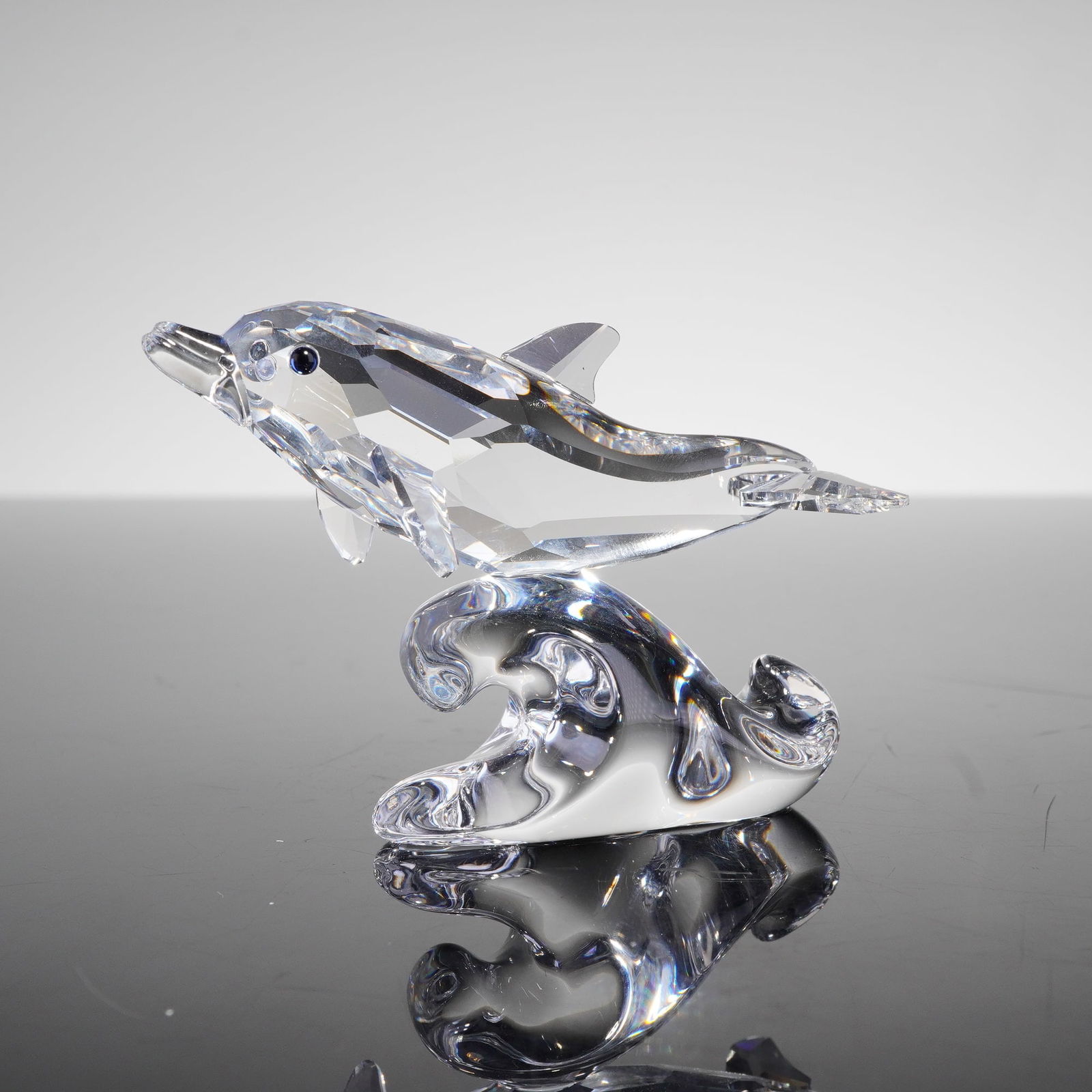 Swarovski Crystal Figurine, Baby Dolphin: A figurine crafted from clear, multi-faceted crystal, depicting a dolphin leaping over a highly polished, stylized crystal wave base. The dolphin features small, blue eyes. The base of the wave elemen