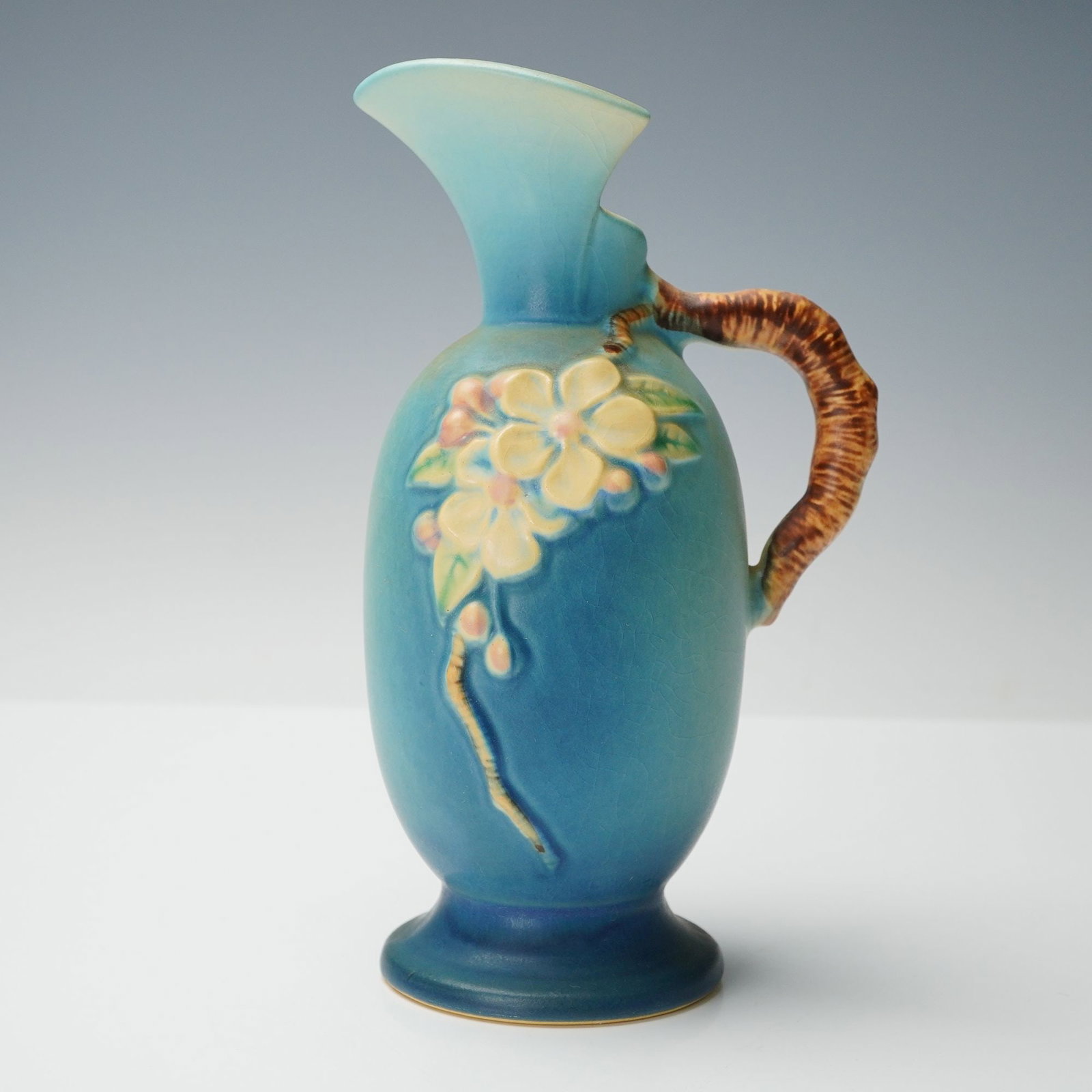 Roseville Pottery Blue Ewer, Apple Blossom: Shaped with an oval body and flaring spout, this ewer shows raised apple blossom branches extending across both sides, finished in blue glaze tones. The ribbed handle is modeled to resemble a branch f