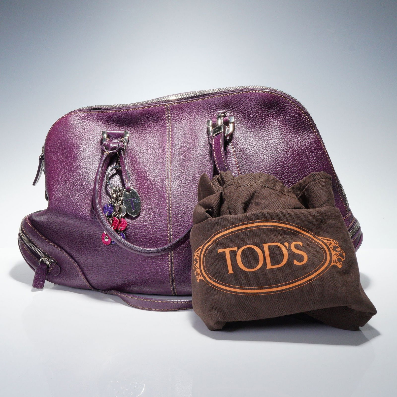 Tods Deep Purple Pebbled Leather Bag: A Tods deep purple pebbled leather bag, featuring a top-handle style with light brown contrast stitching and silver-toned hardware. The bag is designed with double top handles, a main zip closure, and