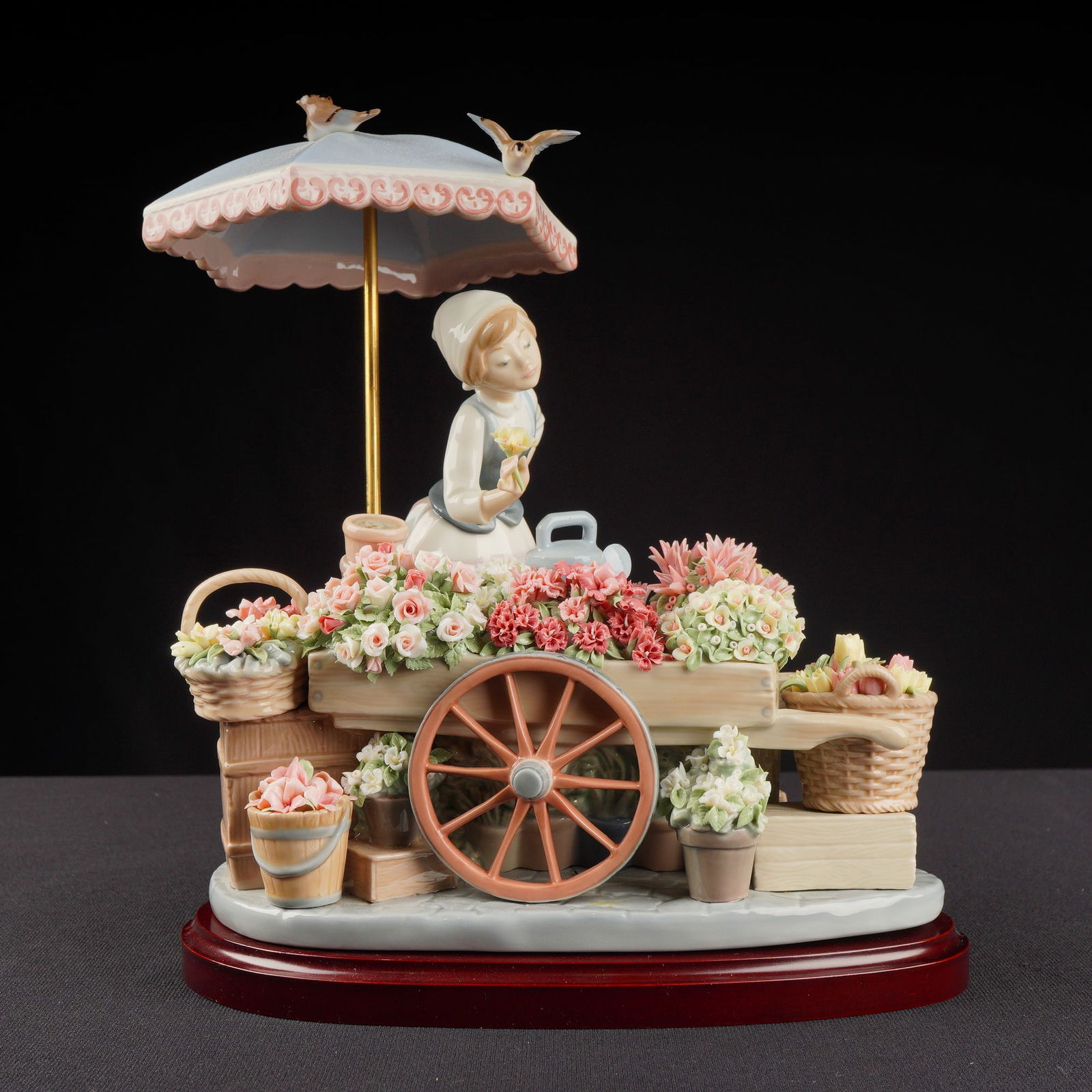 Lladro Porcelain Figurine Flowers of the Season 01001454 with Base: The sculpture depicts a young woman, a flower seller, standing beside a wooden cart laden with numerous finely modeled ceramic flowers and greenery. The scene is sheltered by a light blue parasol supp