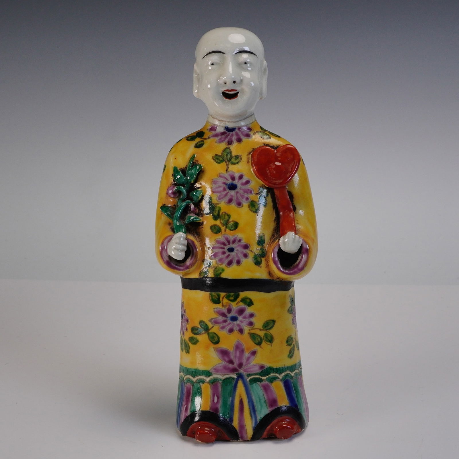 Chinese Yellow Famille Rose Porcelain Joss Stick Holder: The figure is depicted standing and dressed in a long yellow robe decorated with floral and butterfly motifs in green, purple, and pink enamels. He holds a green floral stem in his right hand and a re