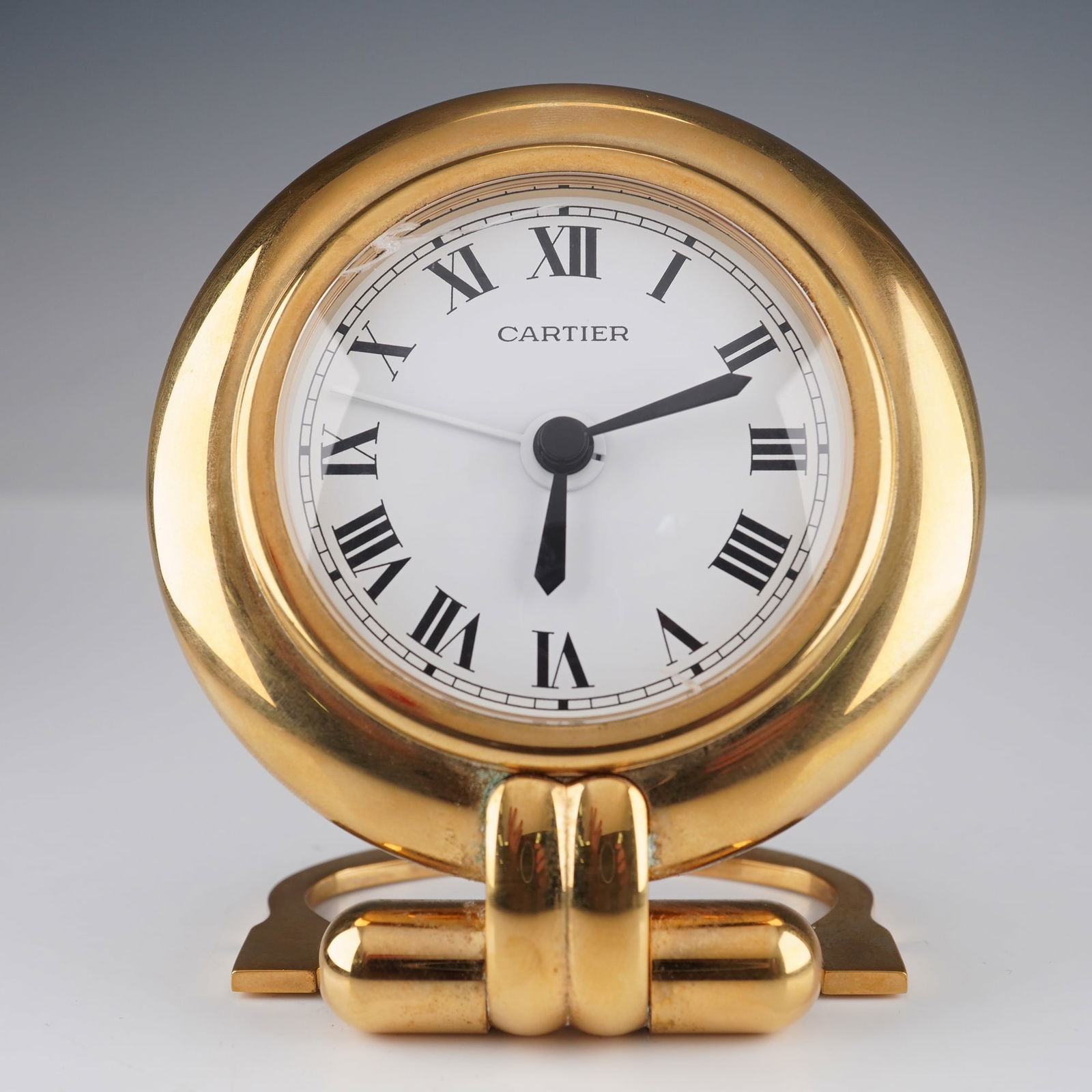 A Cartier Colisee Travel Desk Alarm Clock: A Cartier Colisee Travel Desk Alarm Clock, featuring a circular gold-toned metal case with an integrated folding stand. The front presents a white dial with black Roman numerals, black hour and minute