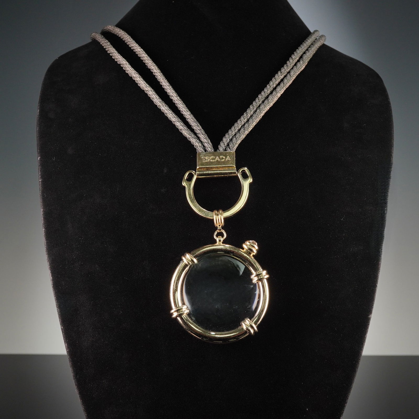 Escada Gold-Toned Magnifier Pendant Necklace with Braided Cord: An Escada Italy fashion necklace featuring a magnifier pendant. The necklace comprises a double-strand, silvery-grey braided metallic cord. The strands are joined by a rectangular gold-toned bar marke
