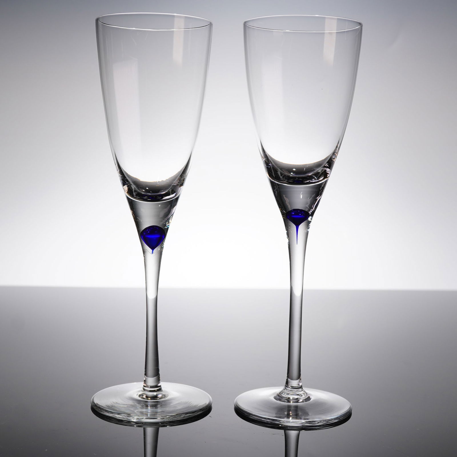 A Pair Crystal Wine Glasses: A pair of stemmed crystal glasses, atributed to Orrefors. Each glass features a tall, tapering bowl suitable for wine, supported by a slender stem and a circular foot. The defining characteristic is t