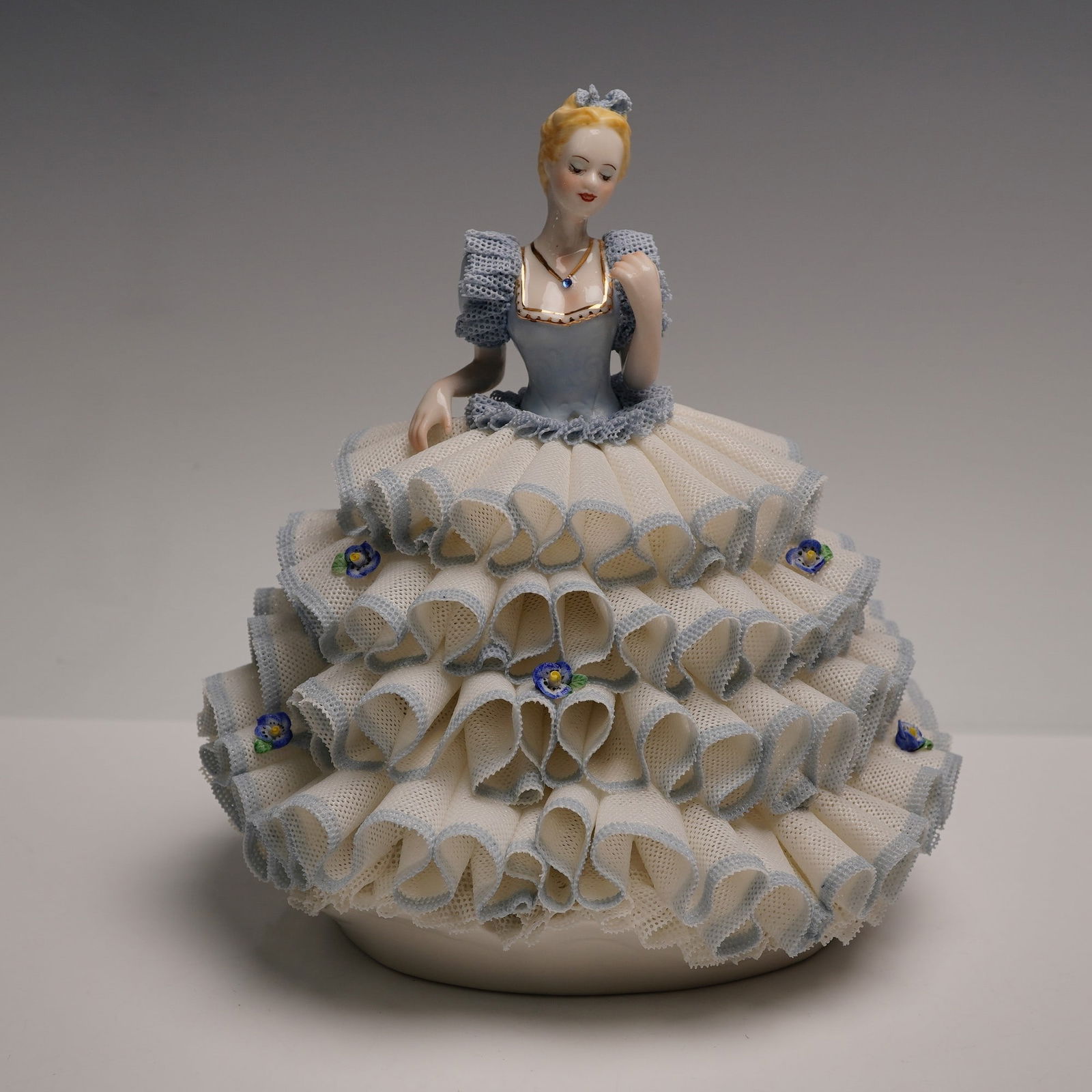 Irish Dresden Lace Porcelain Figurine, Cindy: Charming porcelain figure depicting an elegantly poised young woman adorned in a voluminous tiered gown of intricate lacework accented with delicate blue edging and small applied floral motifs. Painte