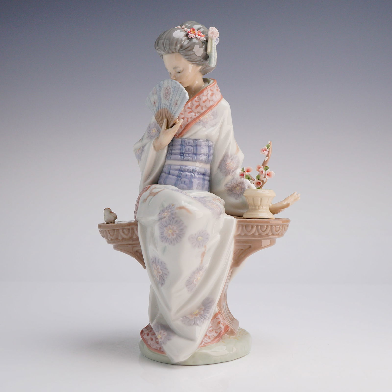 Lladro Porcelain Figurine, Nippon Lady 1005327: A Lladro porcelain figurine, known as the Nippon Lady, model number 5327. This single glazed porcelain sculpture depicts a seated woman, stylized as a Japanese geisha, holding a decorative fan to her