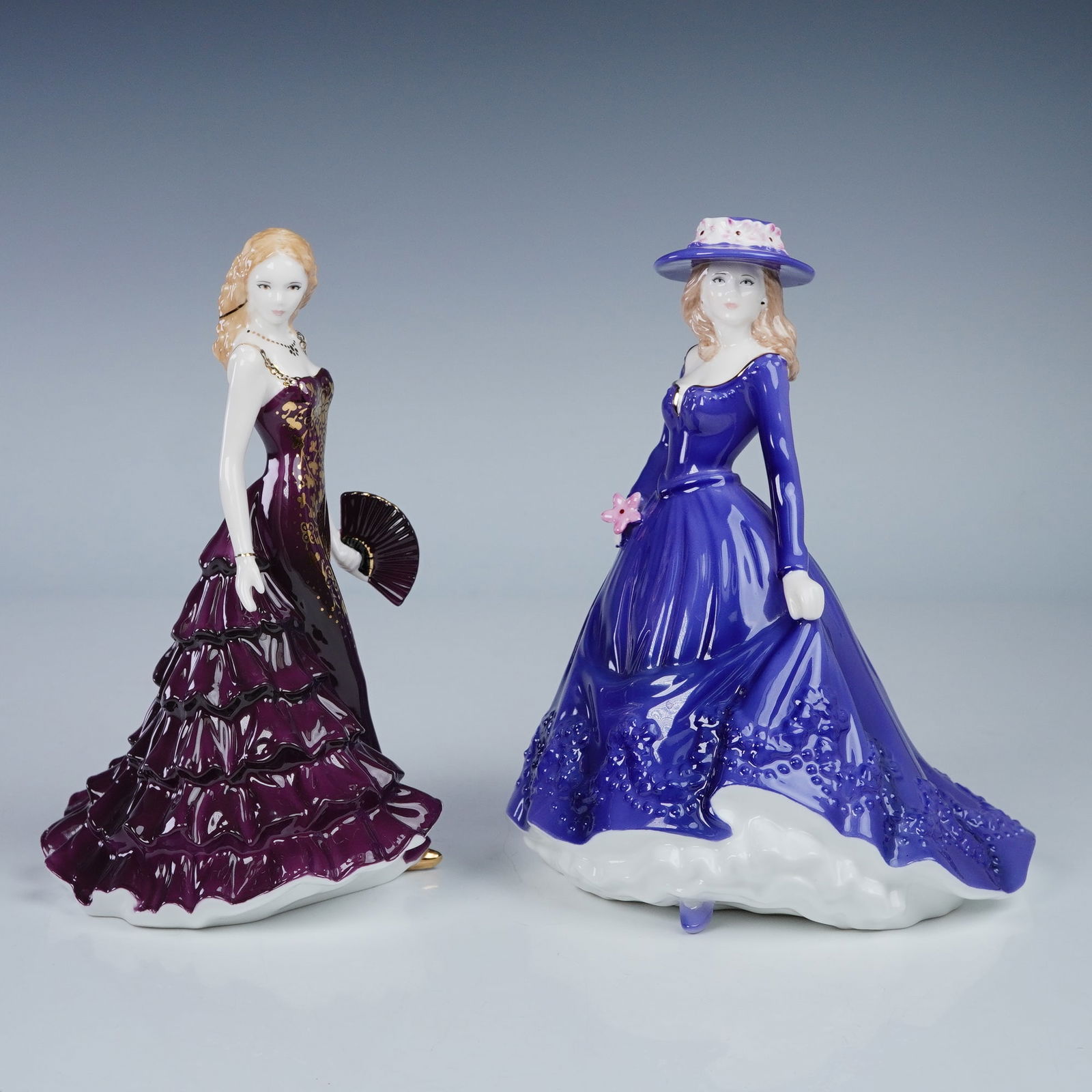 A Pair of Royal Worcester Les Petites Figurines, 'Abigail' and 'Leanne': A pair of Royal Worcester Les Petites ceramic figurines. The left figurine, named 'Abigail,' depicts an elegant woman in a deep plum or burgundy ruffled gown with gold accents, holding a fan. The righ