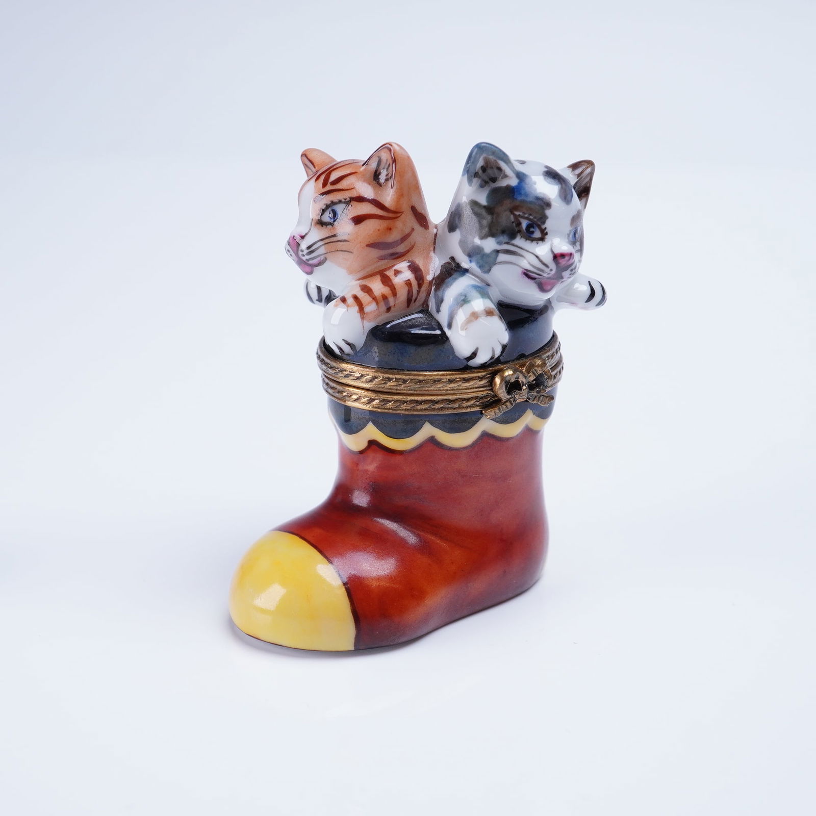 Limoges Trinket Box Kittens in Christmas Boot: This is a hand-painted porcelain hinged trinket box made in Limoges, France titled 'Kittens in Christmas Boot.' The piece features two playful kittens, one orange tabby and one calico, emerging from t