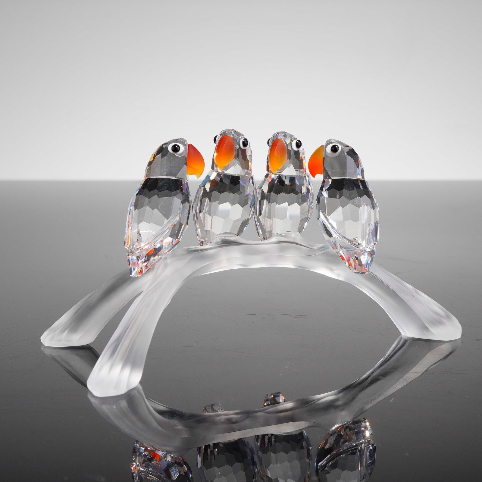 Swarovski Crystal Figurine, Baby Lovebirds: This sculpture consists of four faceted clear crystal birds perched upon a stylized branch constructed of frosted crystal. The birds feature orange beaks and black eyes. The base of the frosted branch