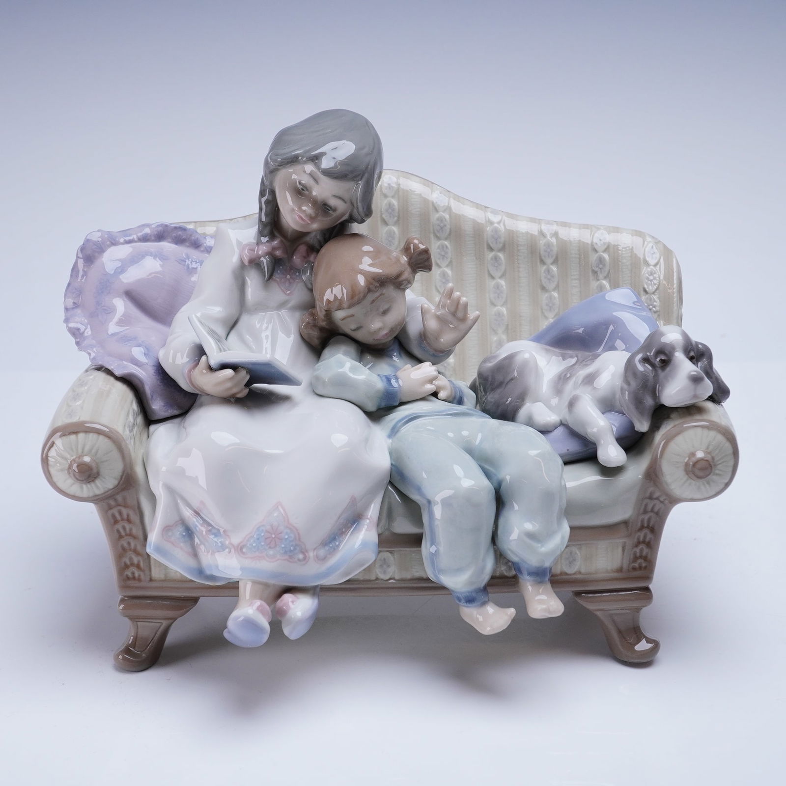 Lladro Porcelain Figurine, Big Sister 01005735: The hand-made glazed porcelain figurine depicts a domestic scene featuring an older girl reading a book while seated on an upholstered sofa. Beside her, a younger child sleeps, resting against her, ac