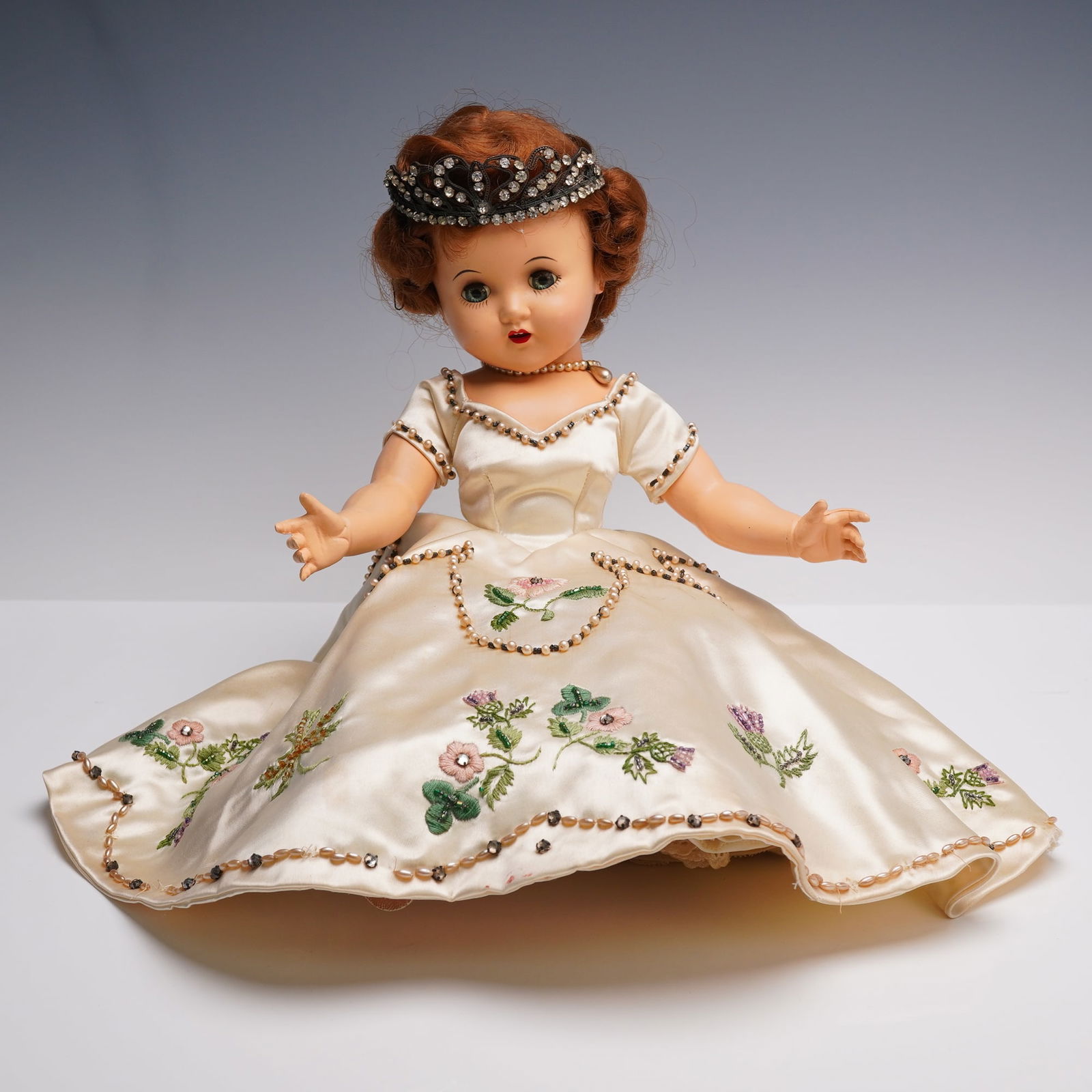 Vintage Hard Plastic Princess Doll in Embroidered Gown with Tiara: A mid 20th century vintage doll dressed as a princess, featuring sleep eyes, red painted lips, and auburn wigged hair styled elegantly. The doll is adorned in a cream satin gown richly embroidered