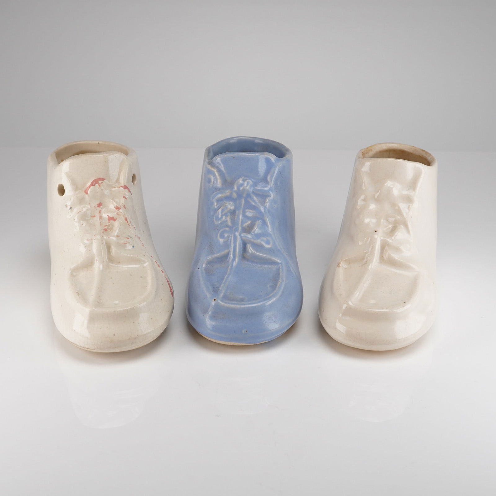 3pc McCoy Ceramic Baby Shoe Planters: A trio of three vintage McCoy ceramic baby shoe planters. The collection includes one light blue shoe situated between two cream or off-white shoes. Each porcelain vessel features molded details