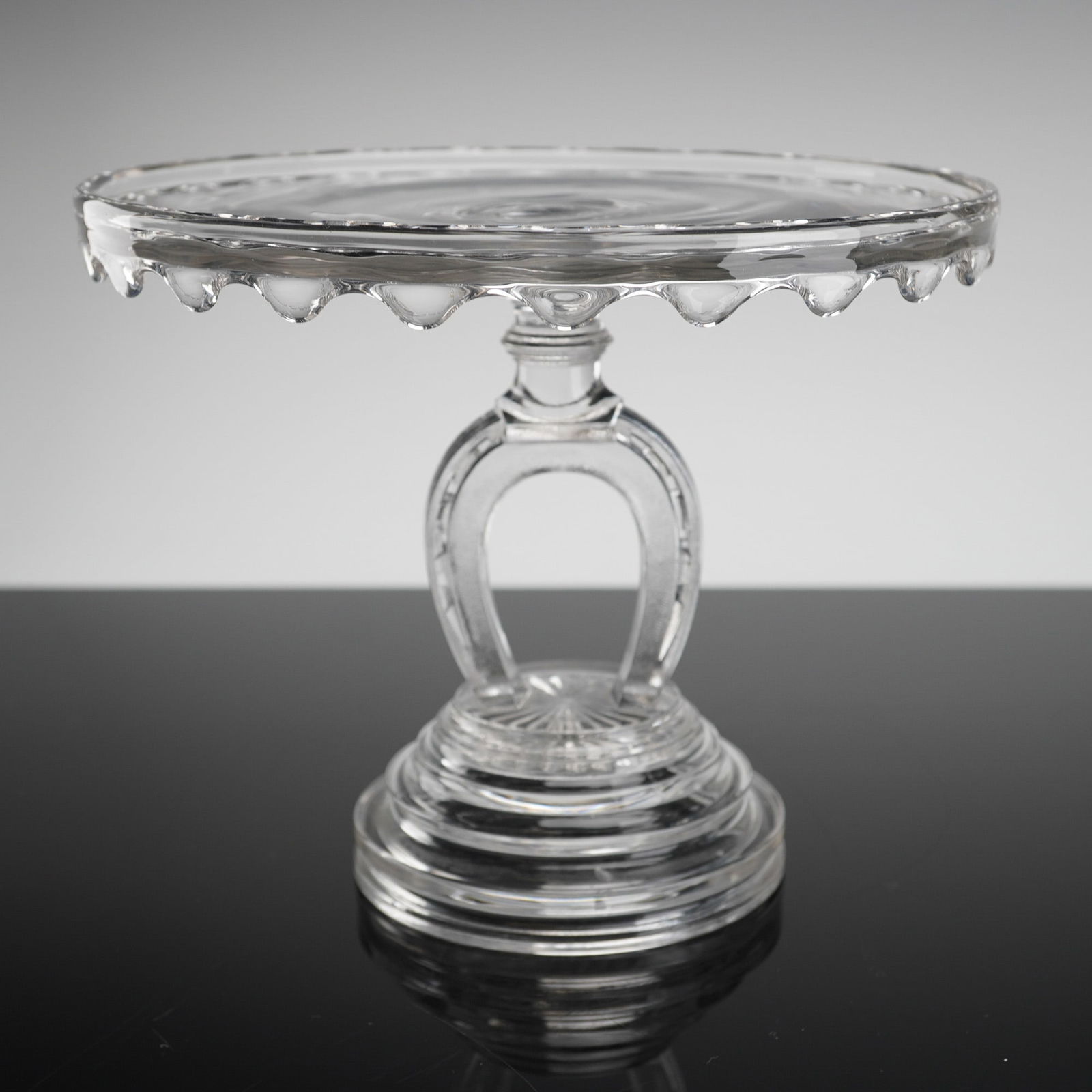 Early American Pressed Glass Cake Stand, Horseshoe Pattern: A single Early American pressed glass cake stand, dating to the late 19th century (Victorian era). The stand is executed in clear, colorless pressed glass, characteristic of the EAPG style. The circul