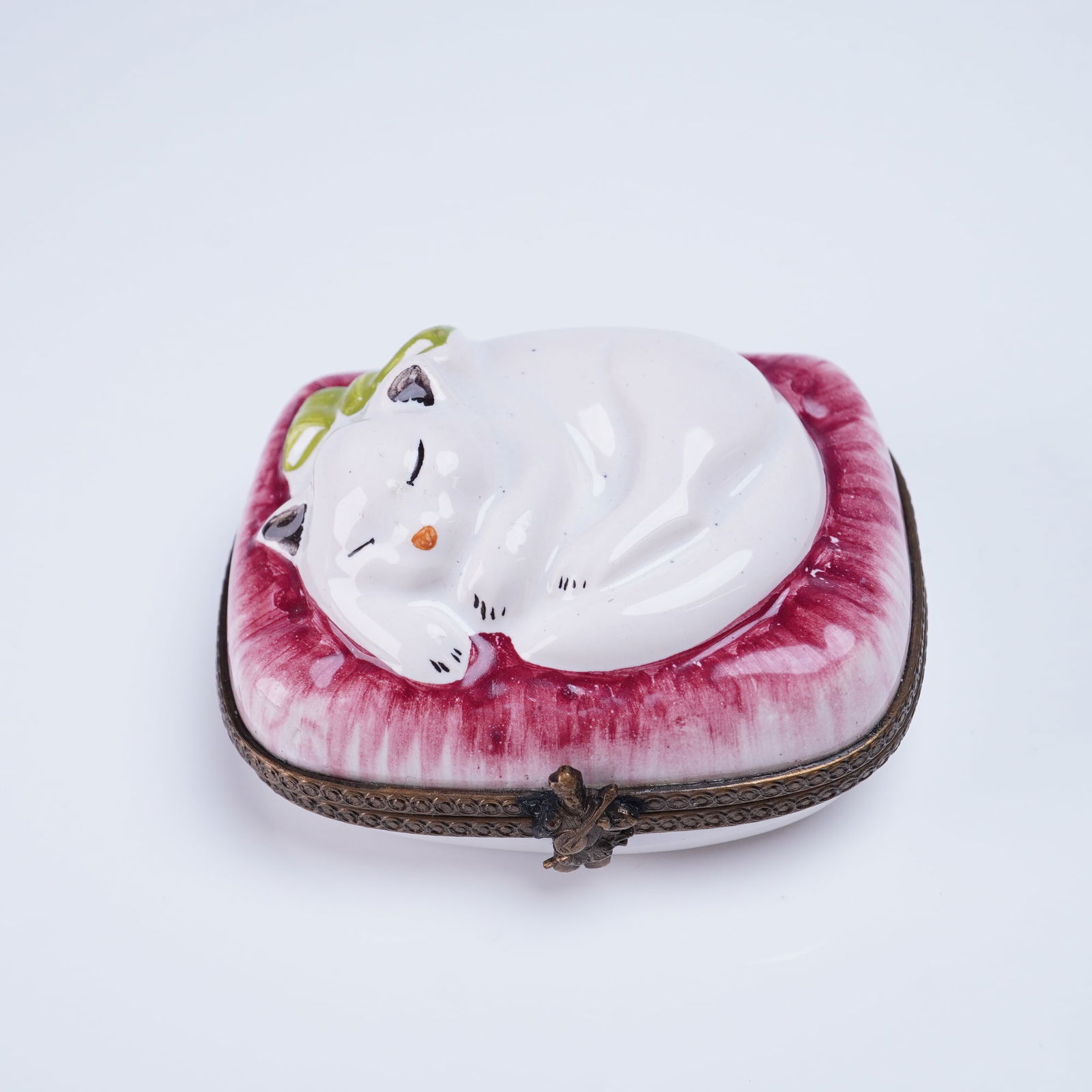 Marshall Fields France Hand-Painted Porcelain Trinket Box Sleeping Cat: A Marshall Field's France hand-painted porcelain hinged trinket box. The rectangular base supports a lid molded and painted to depict a white cat sleeping curled on a textured, deep magenta cushion.