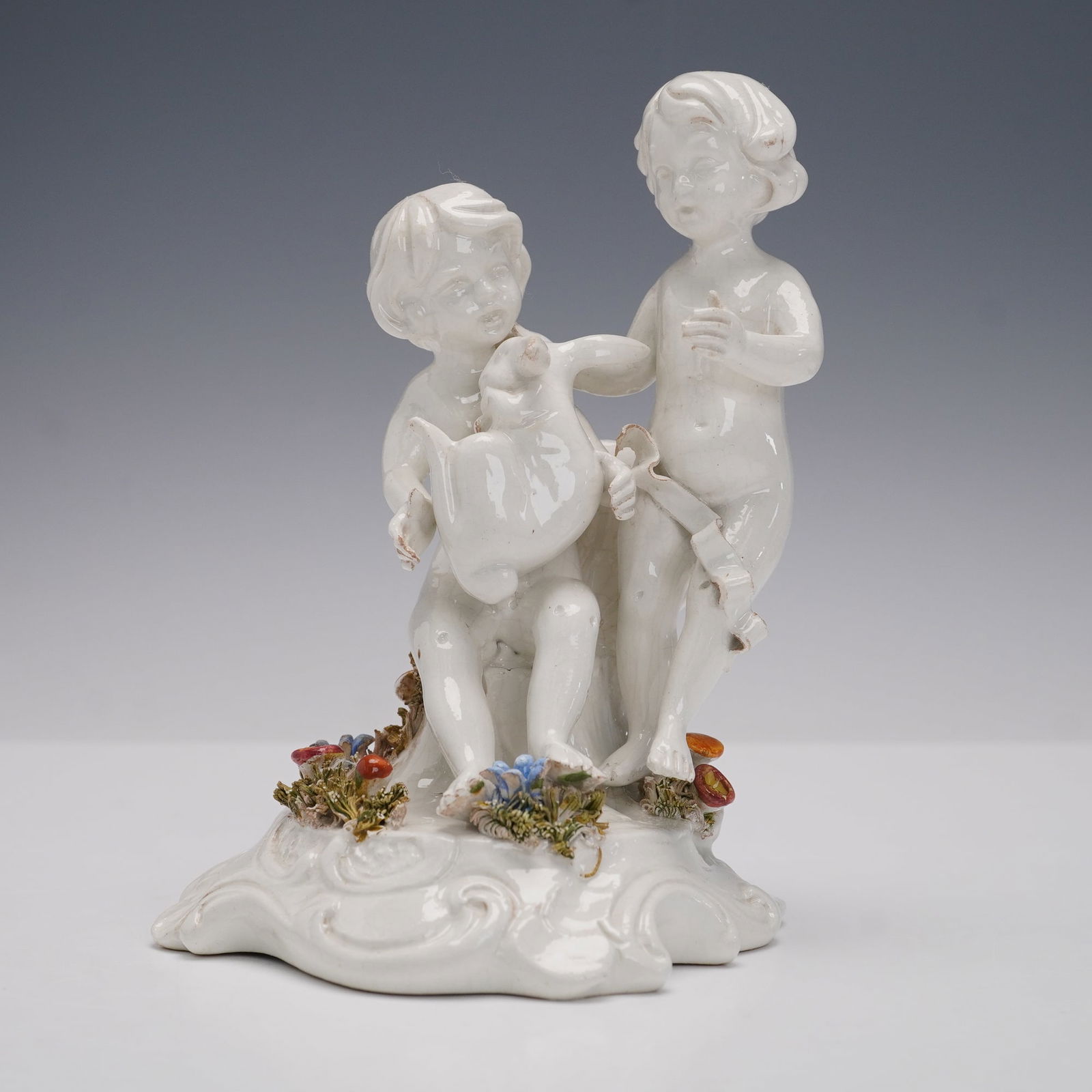 Vintage Porcelain Cherub's Playing Figurine: A pair of cherubs in playful interaction, one holding a rabbit while the other gestures beside him. The figures are set upon a scrolling base accented with applied polychrome flowers and foliage detai