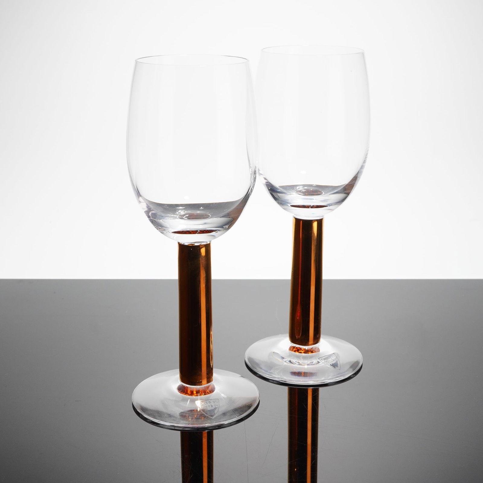 Pair of Orrefors by Gunnar Cyren Nobel Crystal Wine Glasses: Each glass features a clear, glass bowl and a circular foot, connected by a polished, cylindrical stem of copper or bronze-toned metal, refi?ecting a Mid-Century Modern aesthetic. The items are