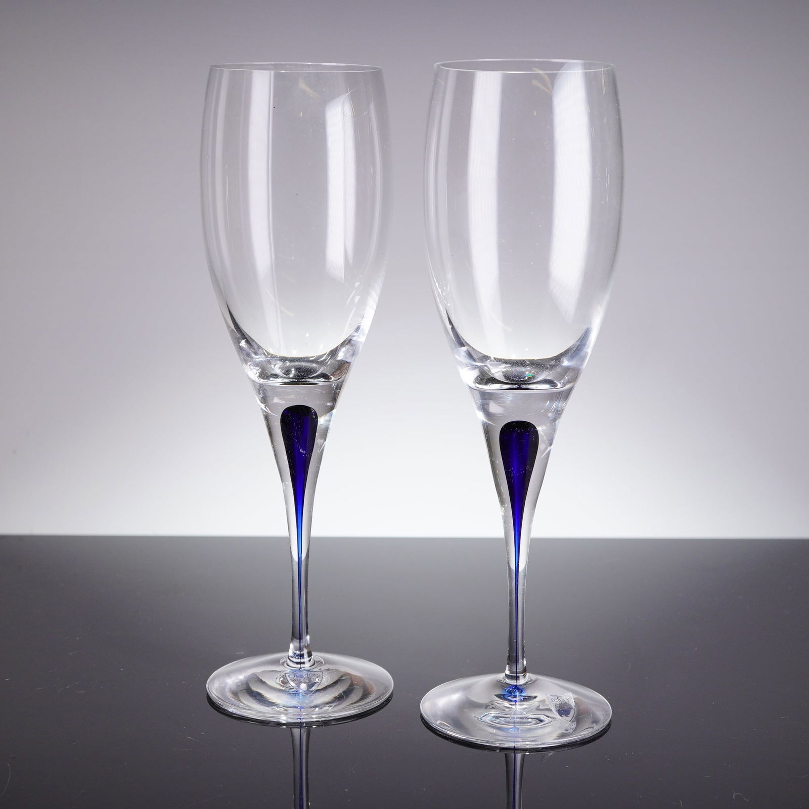 A Pair of Orrefors Intermezzo Blue White Wine Glasses: A pair of contemporary white wine glasses from the Orrefors Intermezzo Blue line, designed by Erika Lagerbielke. Each glass features a clear crystal bowl atop a clear tapering stem. The defining
