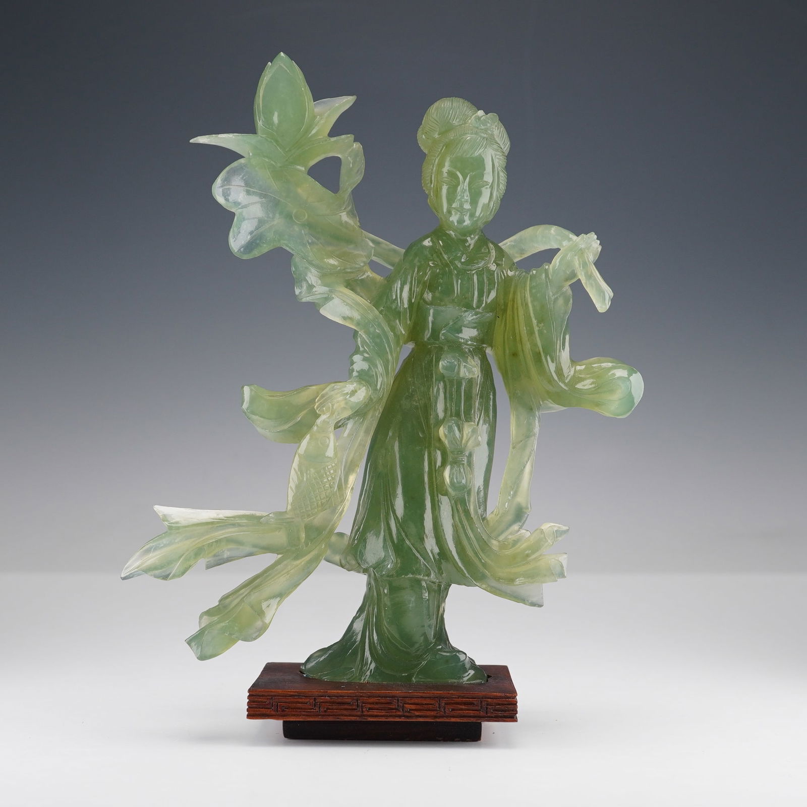 Chinese Carved Green Jade Figurine, Guanyin: Depicts a standing female figure in traditional style, characterized by her elaborate coiffure and gracefully flowing robes and ribbons that extend dynamically around her form. The figure is mounted