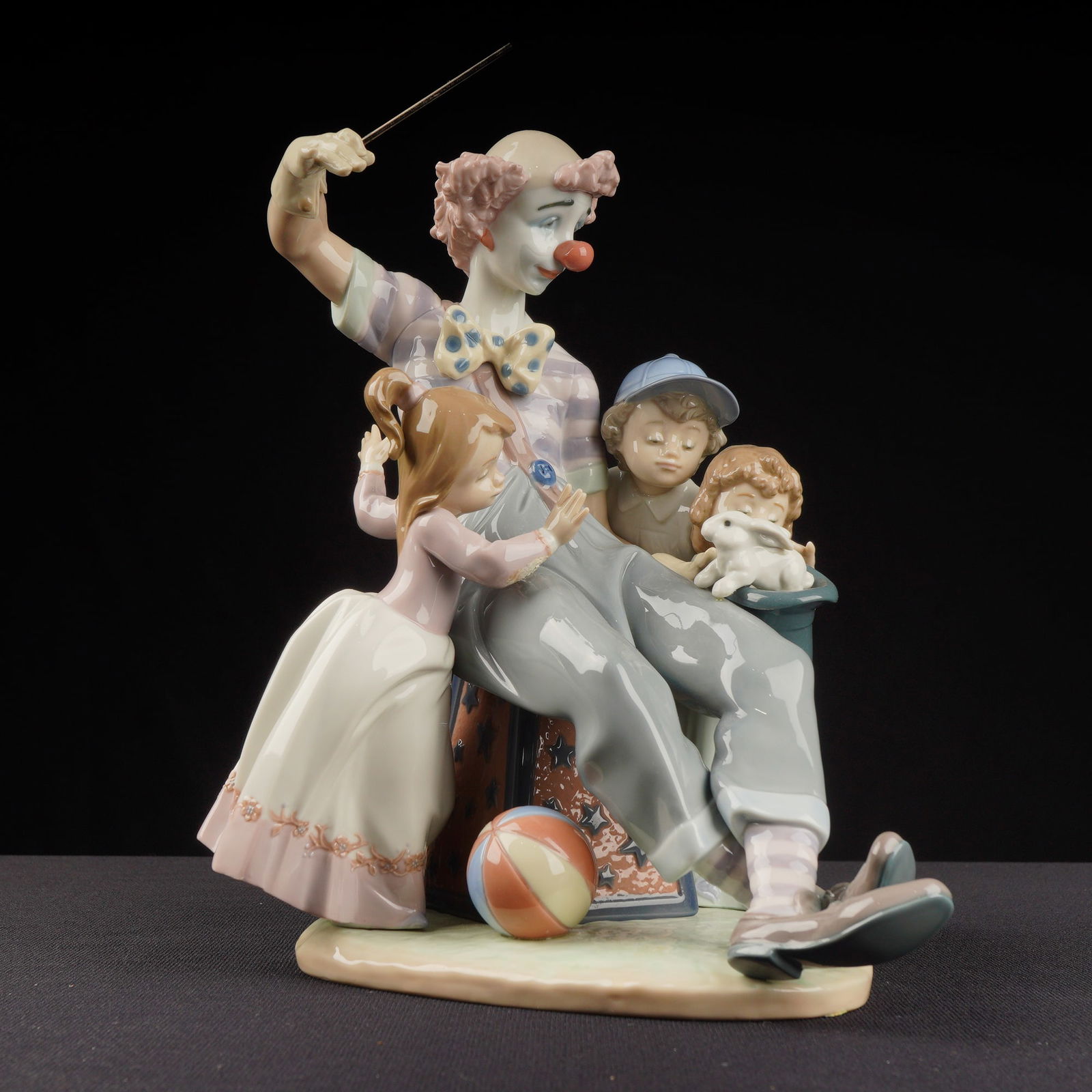 Lladro Porcelain Figurine Magic of Laughter 01005771: The Lladro Porcelain Figurine depicts a contemporary scene of a clown, wearing a striped shirt and large pants, seated on a star-and-moon decorated pedestal while presenting a magic trick, surrounded