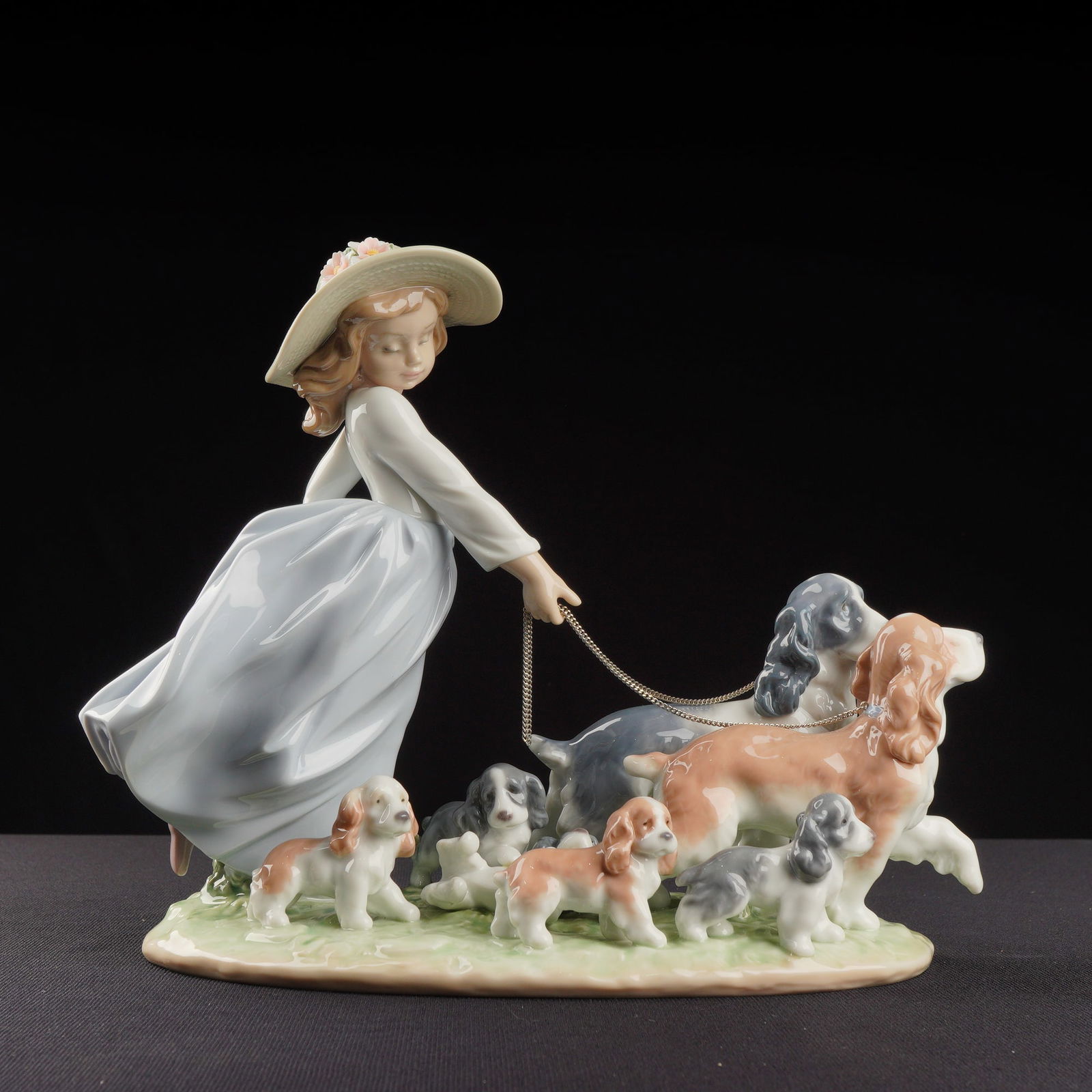 Lladro Porcelain Figurine Puppy Parade 01006784: This glazed porcelain figurine group depicts a young woman in a flowing white and light blue dress and a straw hat adorned with flowers, walking seven individually sculpted spaniel dogs of various