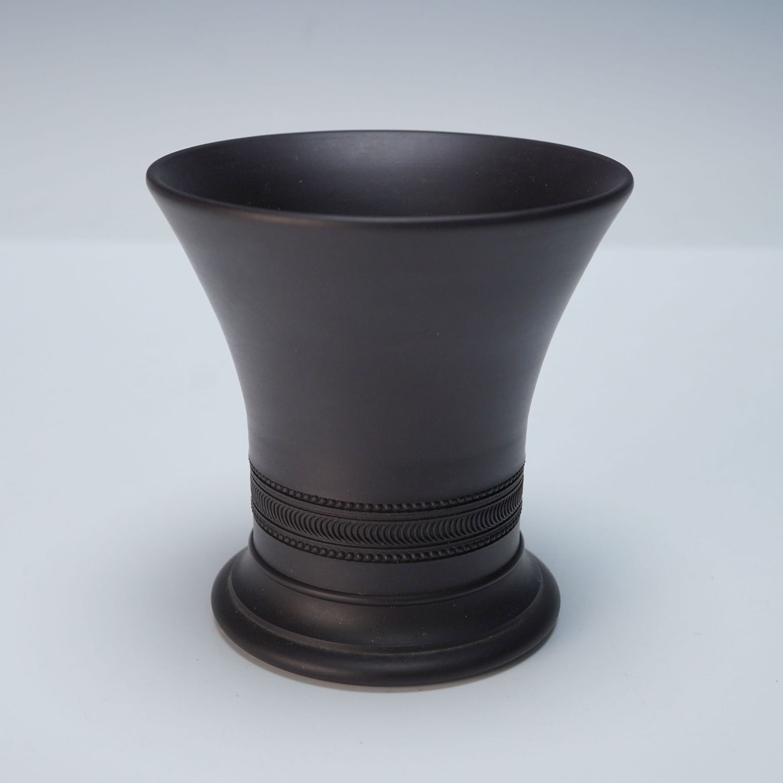 Wedgwood Black Basalt Vase, Signed E. A. Wedgwood: A matte, unglazed ceramic vessel with a flaring rim, concave body, and circular foot. The lower body is encircled by a repeating decorative band of semi-circular motifs and dots. The base is impressed
