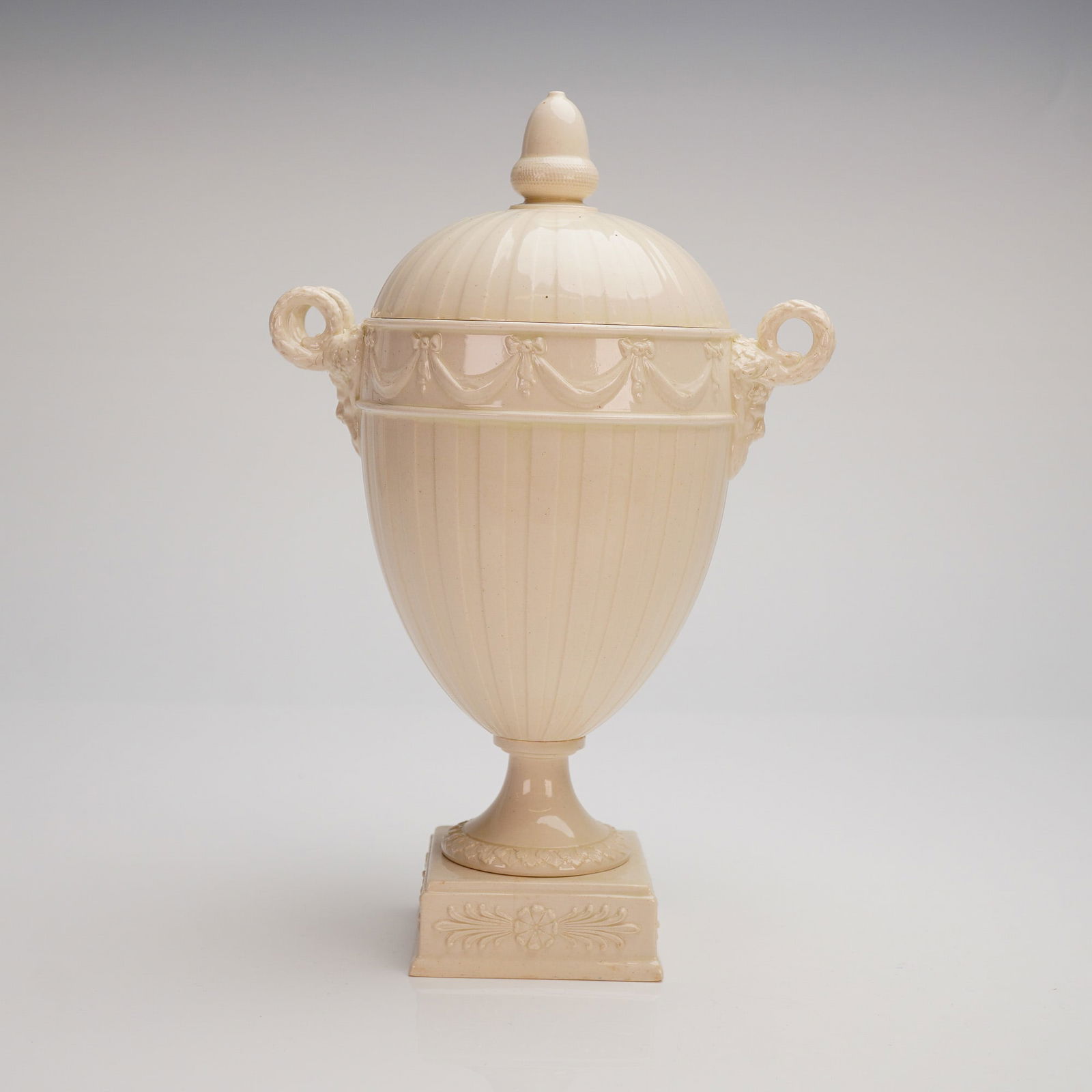 Wedgwood Creamware Satyr-Handle Lidded Urn: The urn features an ovoid body topped with a domed lid, each embellished with vertical fluting. It rests on a square pedestal base decorated with a subtle low-relief foliate motif. The shoulder is enc