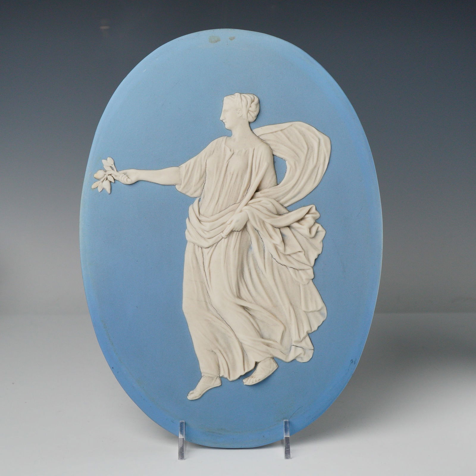 Wedgwood Blue Jasperware Classical Figure Oval Plaque: An oval Wedgwood Blue Jasperware ceramic plaque featuring a white high-relief bas-relief classical female figure set against a pale blue ground. The figure is depicted in profile, dressed in flowing c