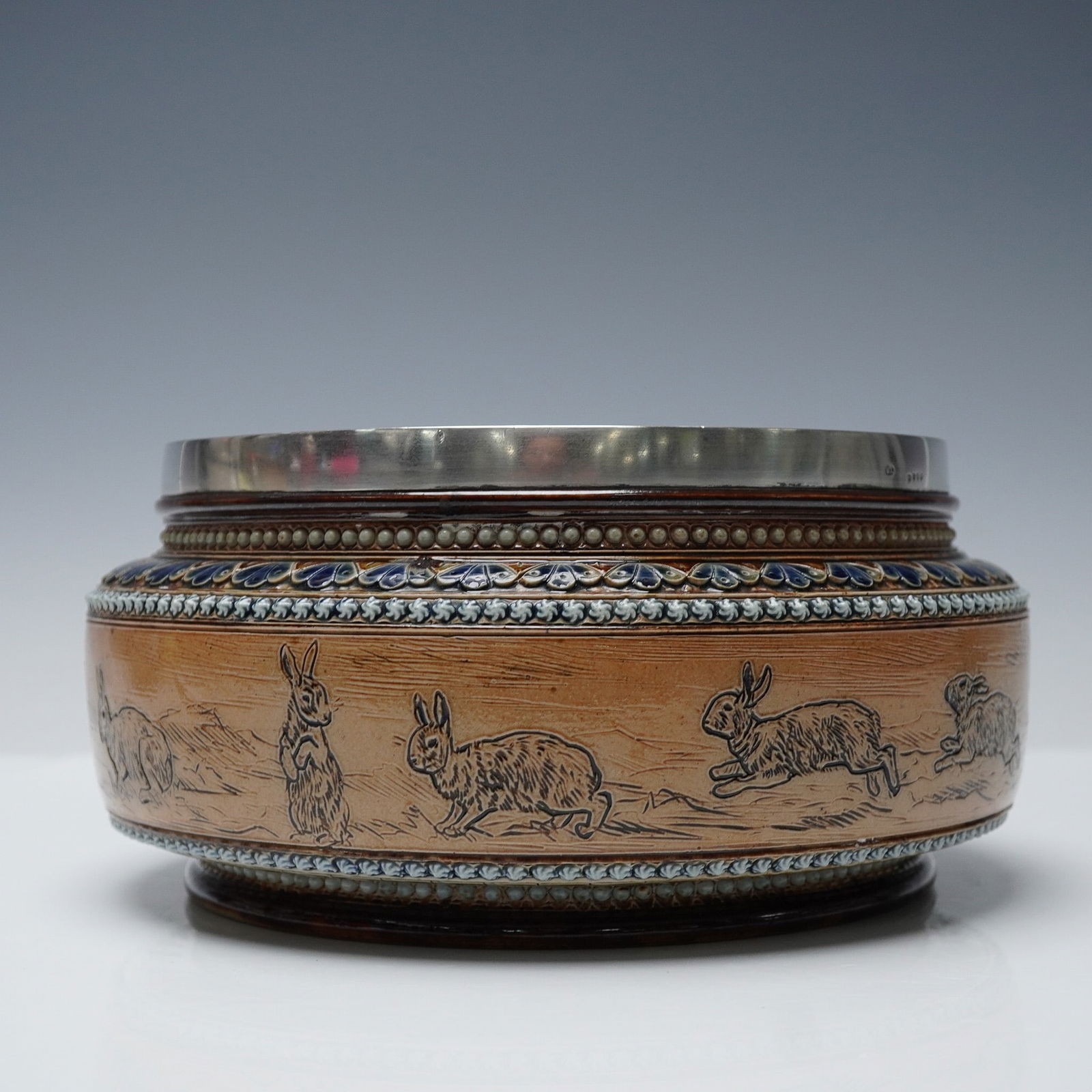 Doulton Lambeth Hannah Barlow Stoneware Sterling Silver Rabbit Bowl: Royal Doulton stoneware bowl featuring an incised continuous scene of rabbits in various poses, designed and executed by Hannah Barlow. The piece is decorated with a raised Art Nouveau foliate border