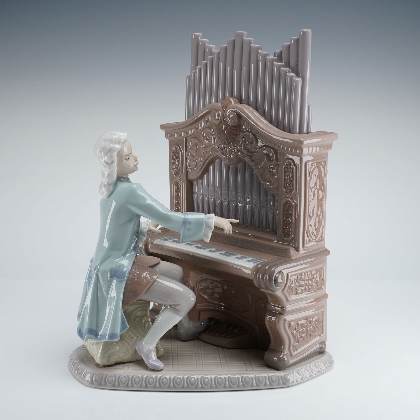 Lladro Young Bach Sculpture 1001801: The glazed porcelain group depicts a male musician, likely J.S. Bach, seated on a stool and playing an elaborately decorated brown pipe organ featuring gray/lavender pipes. The musician wears