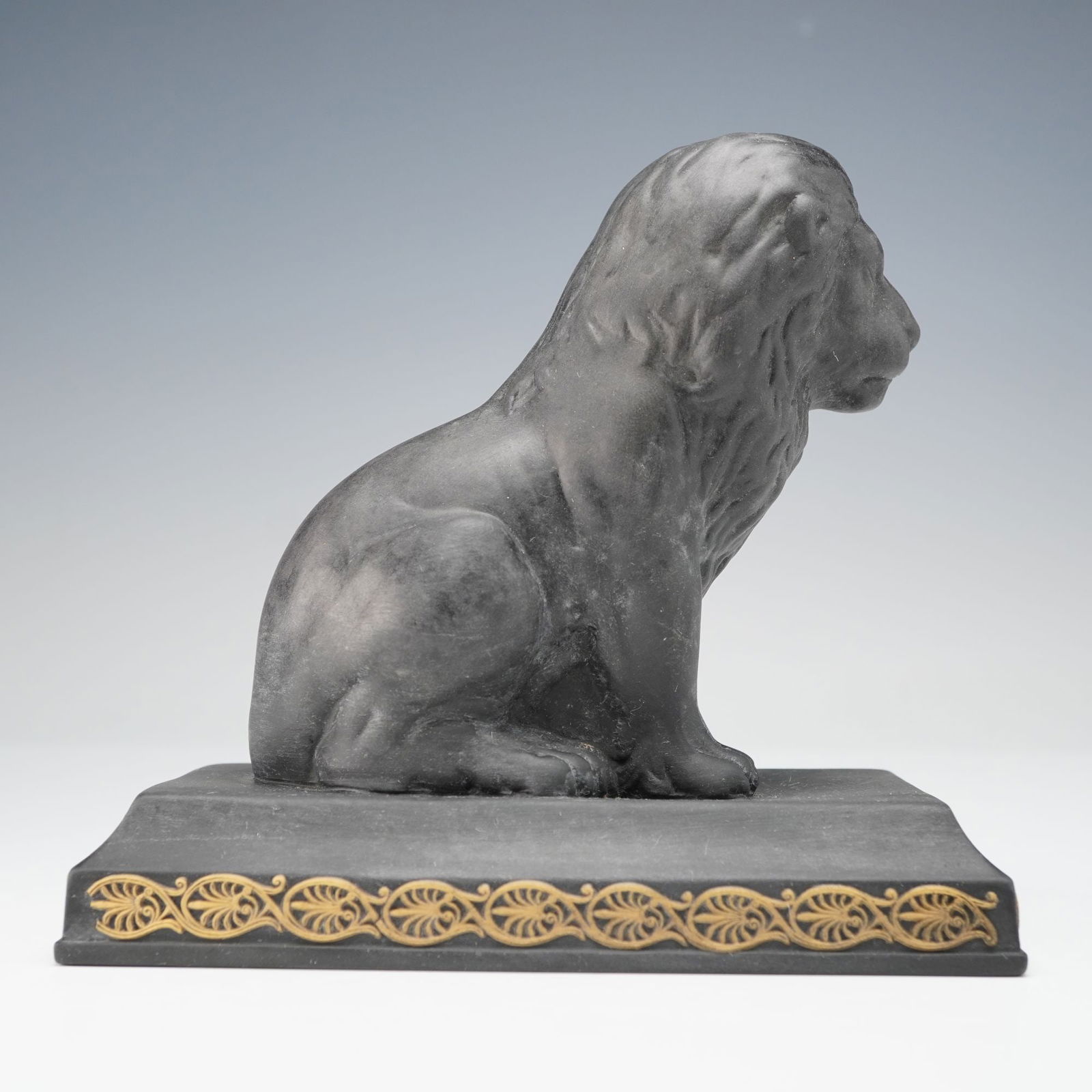 Wedgwood Black Basalt Seated Lion Figurine: Crafted from dark, matte black basalt, depicting a classically styled, seated lion resting on a rectangular plinth base. The base is embellished with a gilded acanthus leaf frieze, reflecting a