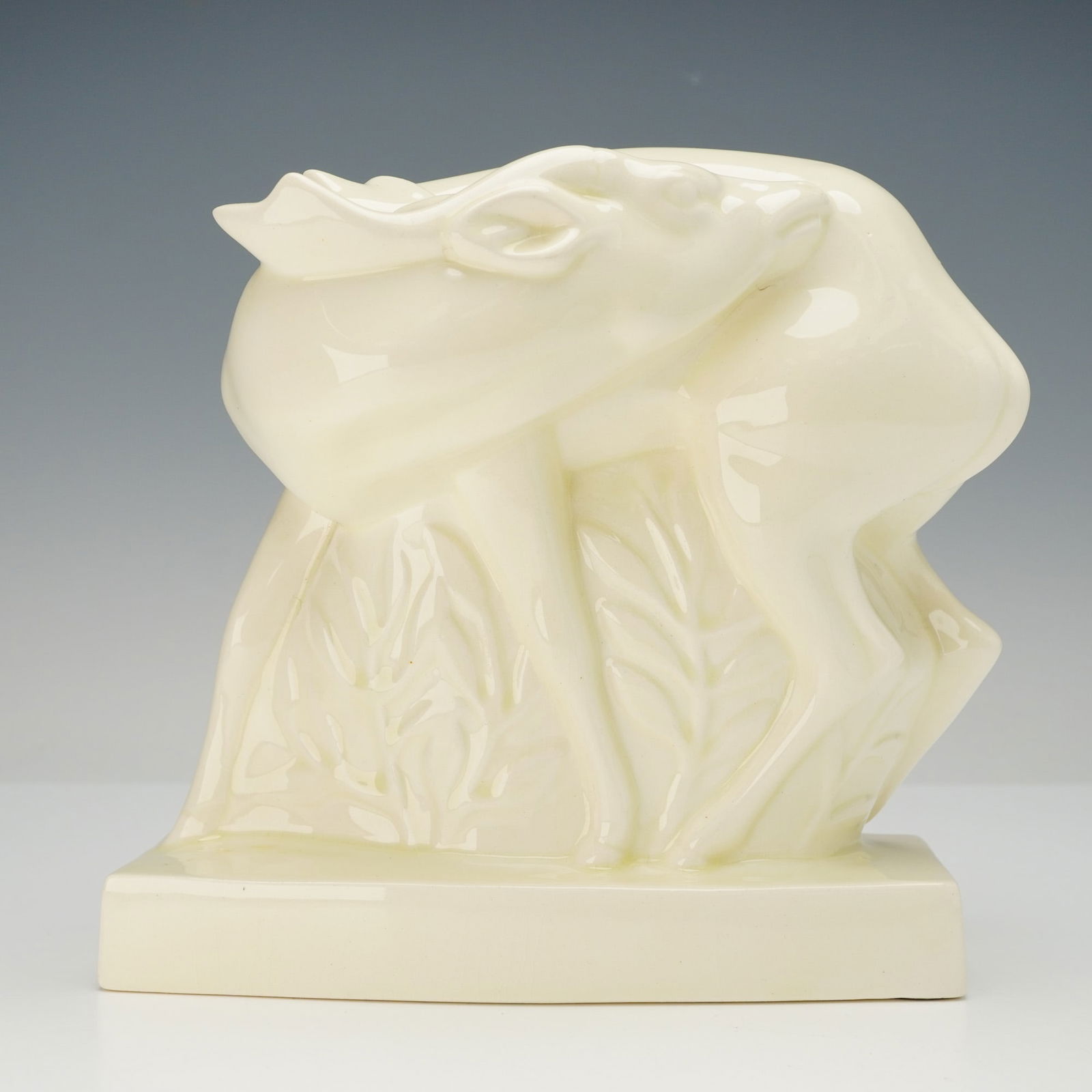 A Wedgwood Art Deco Fallow Deer Figure by John Skeaping: A glazed ceramic figure of a fallow deer, designed by John Skeaping for Wedgwood, embodying the Art Deco aesthetic. The stylized deer is depicted standing amidst foliage, presented in a creamy white,