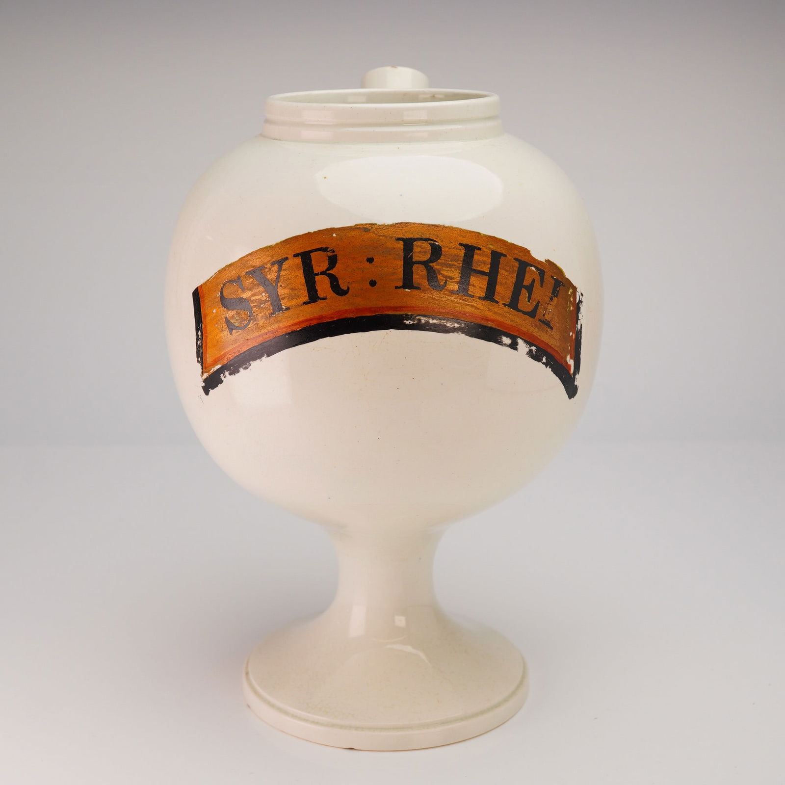 Wedgwood Ceramic Apothecary Jug, Syr: Rhei: Comprising a spherical body set on a tall pedestal base with a flared foot, this cream-glazed vessel features a short cylindrical spout emerging from the shoulder and an open neck, indicating loss of
