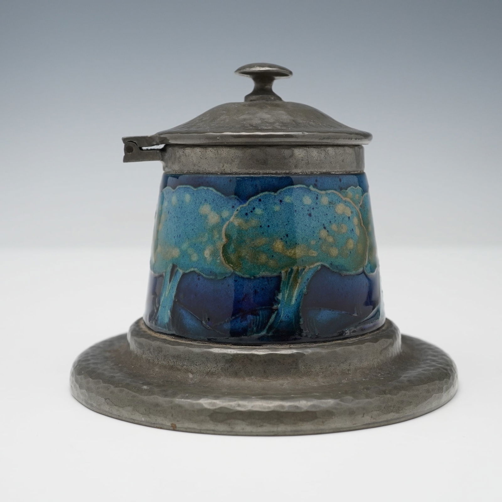 Moorcroft Pottery For Liberty & Co. Tudric Pewter Inkwell, Moonlit Blue: A inkwell comprising a Tudric hammered pewter base and hinged lid, enclosing a tube lined decorated holder. Depicts a band of stylized trees against a cobalt and indigo ground. Tudric Moorcroft and 01