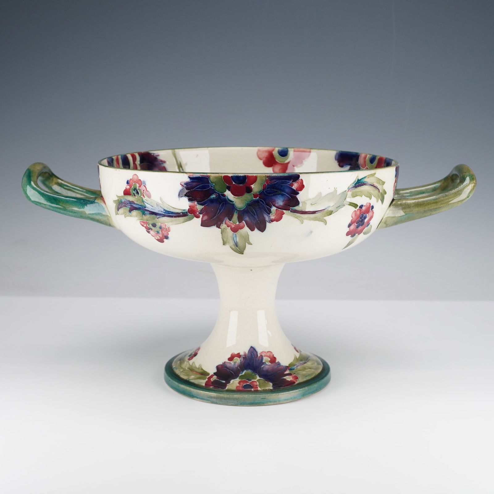 William Moorcroft Pottery Handled Footed Bowl, Persian: An early 20th century bowl of twin handle form with stylized floral and foliate motifs in rich hues of cobalt blue, crimson, and verdant green upon a cream ground. William Moorcroft signed. "M 67"