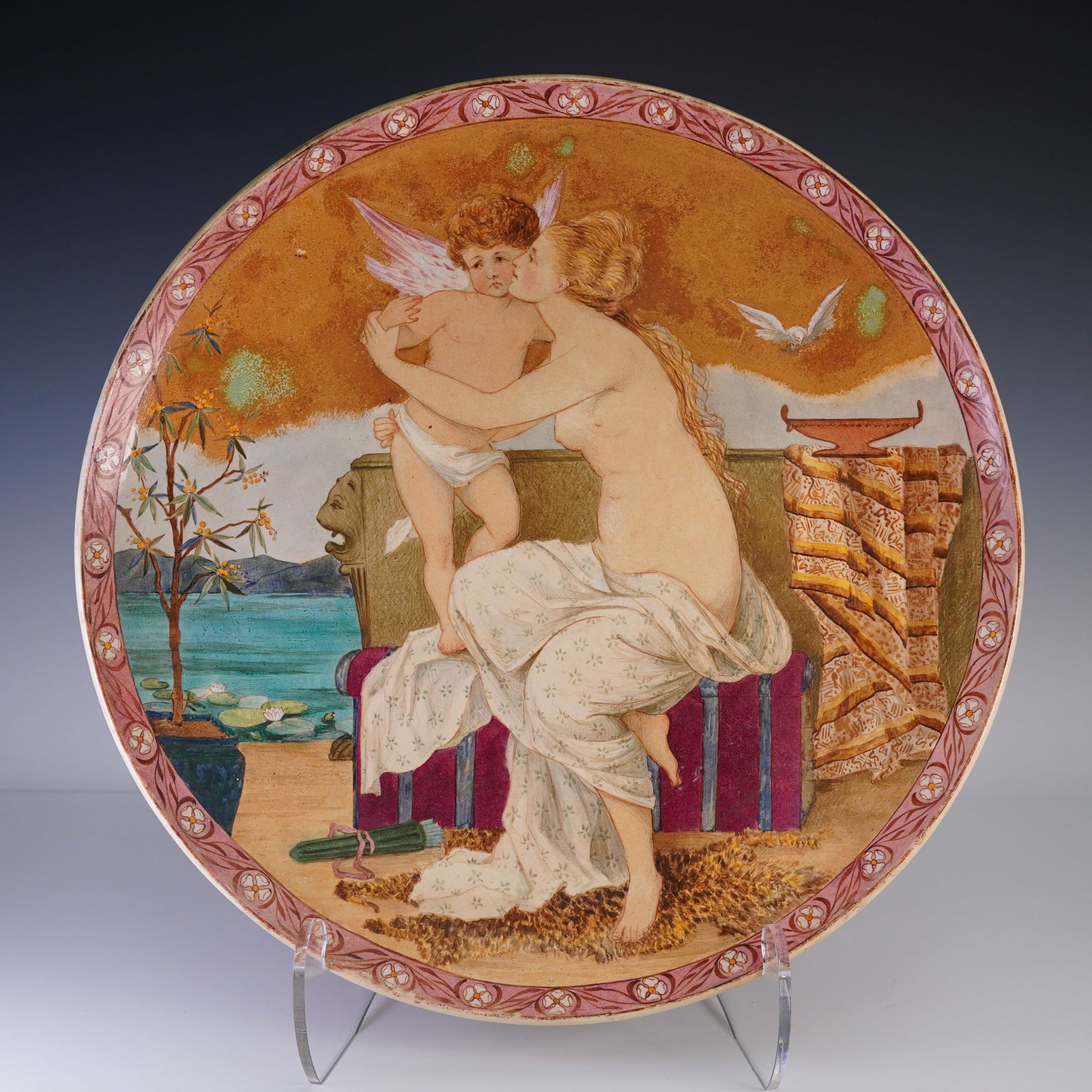 Minton Faience Classical Scene Wall Charger: Hand painted, large earthenware wall charger decorated with a classical scene depicting a seated nude woman kissing a cherub on the cheek. Set on a stylized, daylight scene in warm hues of brown, blue