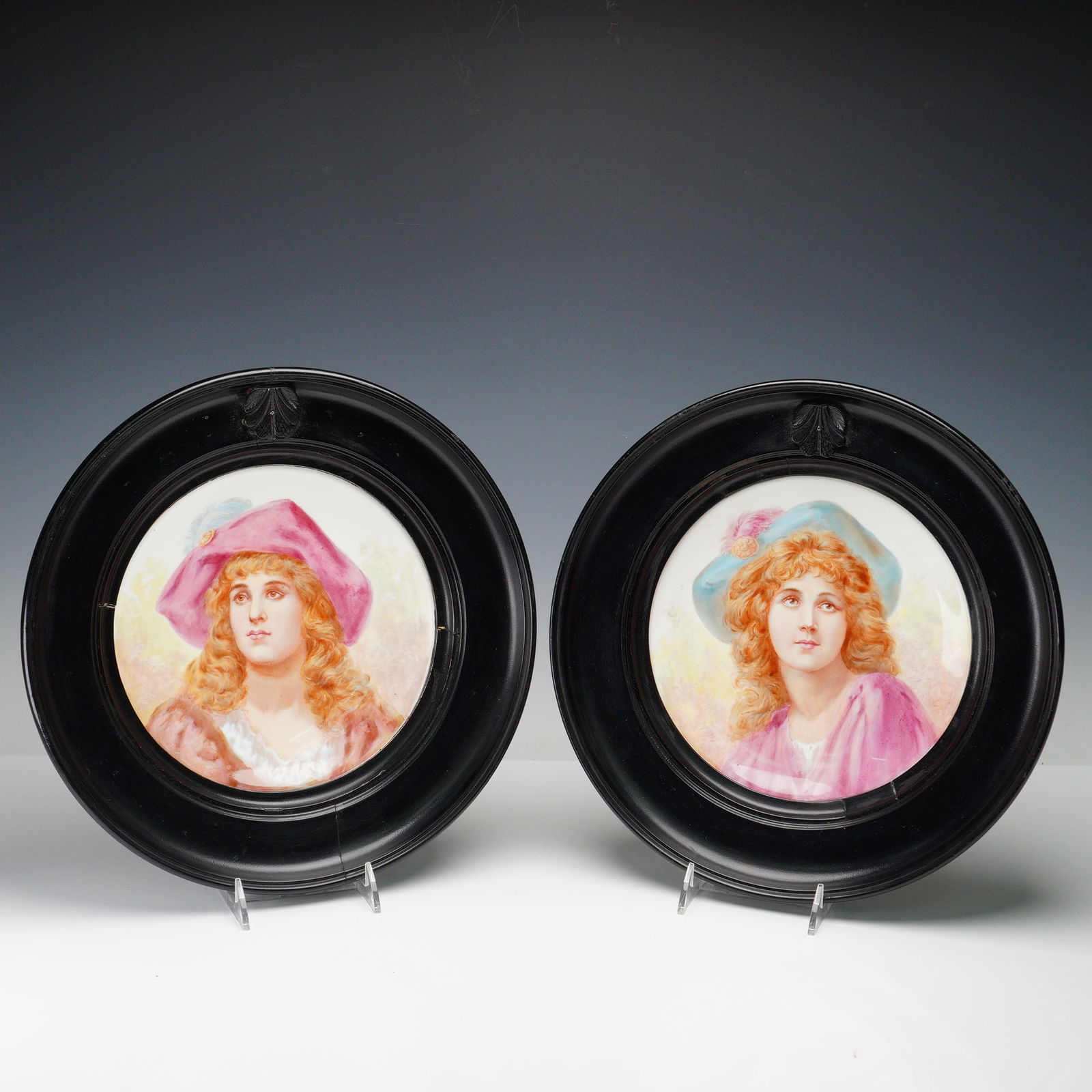 Pair of Artist-Painted Porcelain Plates Framed by J.P. Hewitt: The plates feature portraits of a woman and a man. The woman is wearing a blue hat with a feather and a more modest shirt. The man is dressed in a pink hat, and both figures have soft, flowing hair.