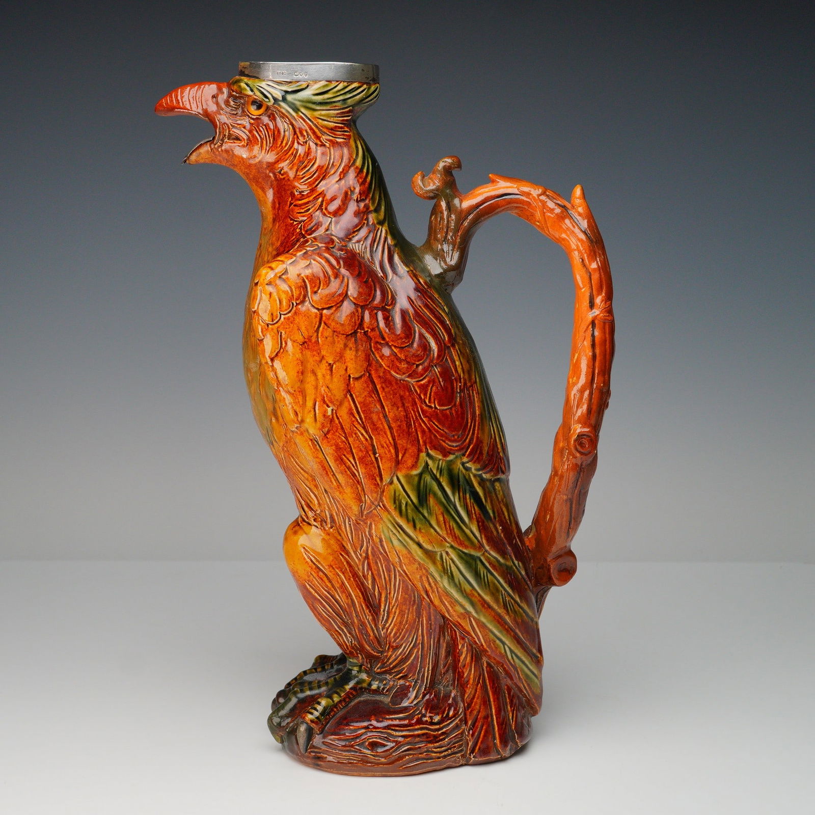 Doulton Lambeth Stoneware and Sterling Silver Decorative Eagle Jug: Figural jug modeled in the form of a perched eagle with finely detailed plumage, talons, and beak, rendered in richly colored stoneware with deep russet, green, and ochre glaze. The eagle's beak and