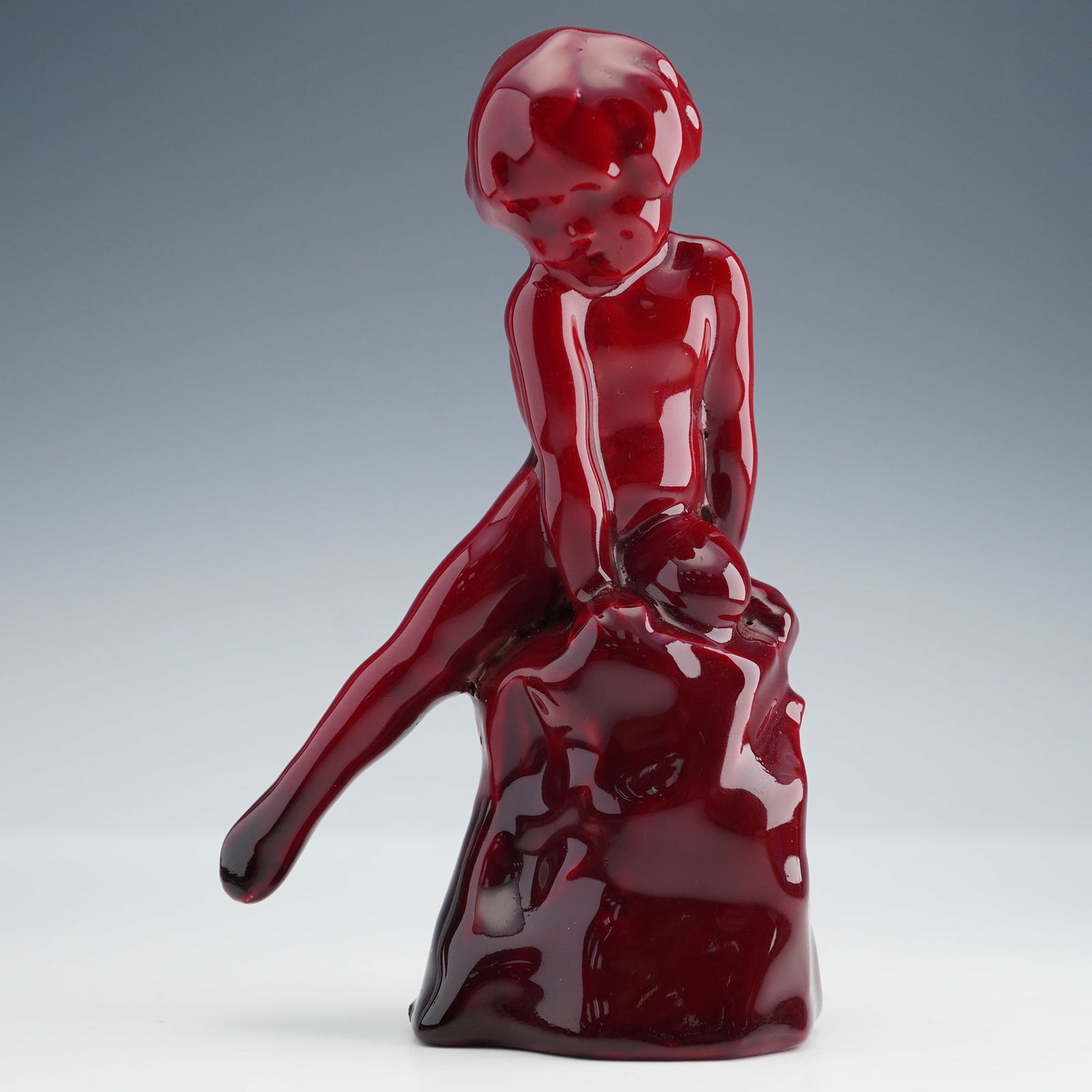 Rare Royal Doulton Red Flambe Figurine, Child on Rock HN603: A striking Royal Doulton Flambe figurine titled Child Study HN603 depicting a young child seated with one leg extended. This piece is rendered in a deep rich flambe glaze that highlights the sculptura