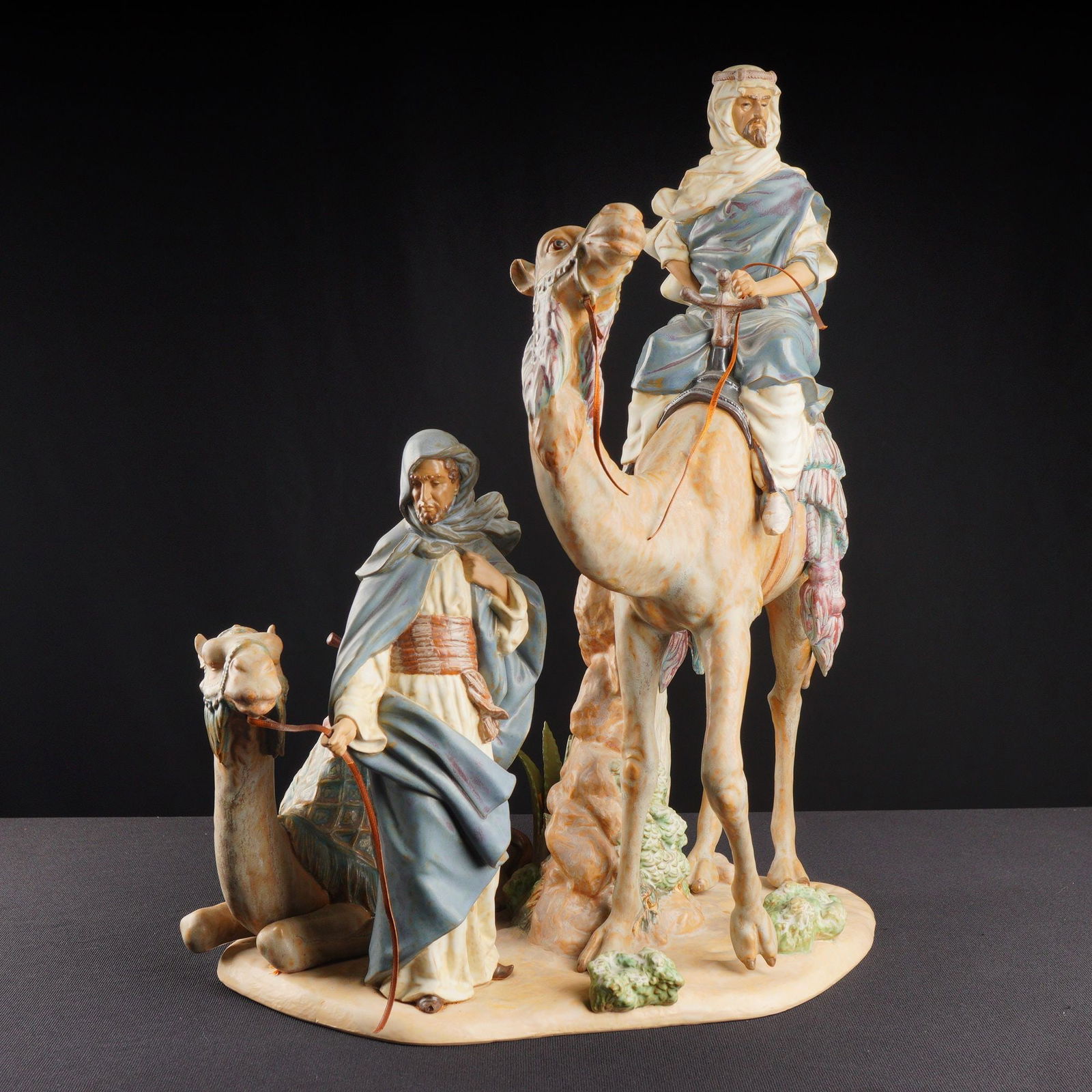Lladro Matte Sculpture Desert People 01013555: The large porcelain figurine depicts a desert scene featuring two figures in robes, one seated atop a standing camel and the other walking beside a resting camel. The piece utilizes a mixed finish of