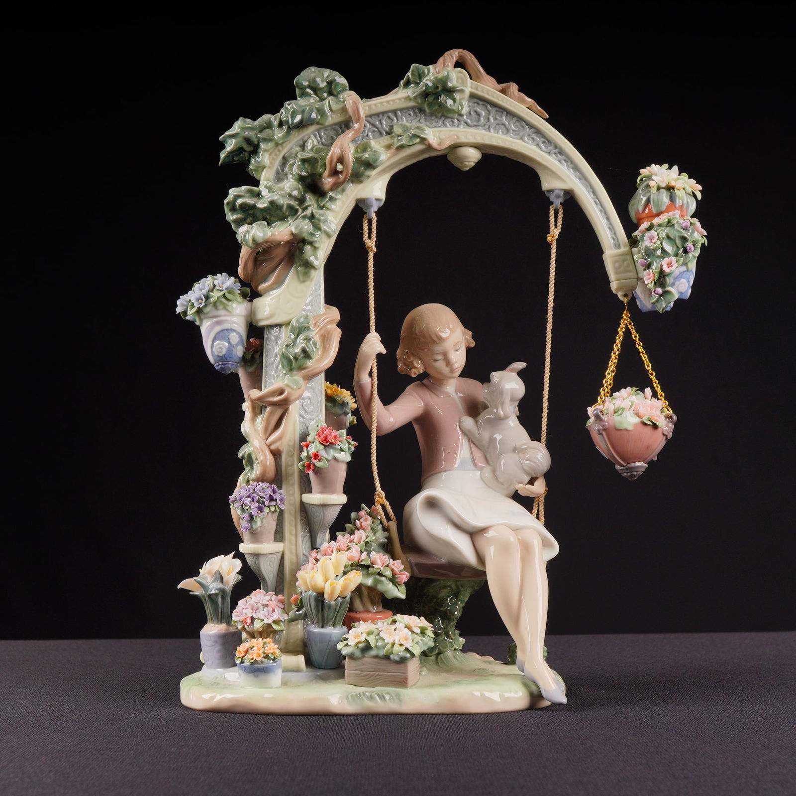 Lladro Porcelain Figurine Swing into Spring 01008311: A large, glazed Lladro porcelain figurine titled "Swing into Spring," model number 01008311. The sculpture depicts a young girl in pastel attire seated on a swing suspended within an elaborate,