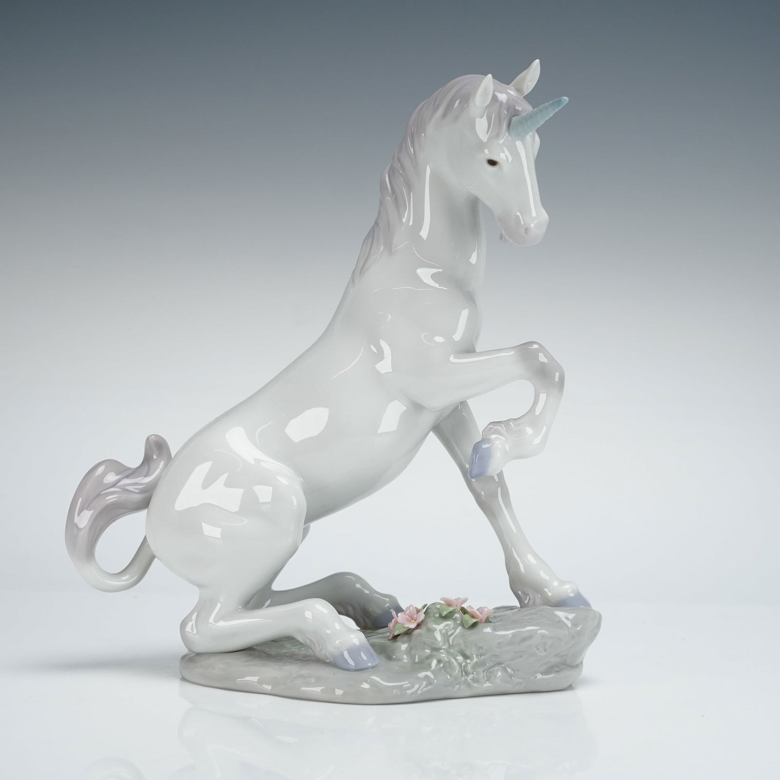 Lladro Porcelain Figurine Magical Unicorn 01007697: The piece is executed in glossy porcelain and depicts a unicorn in a semi-kneeling, rearing pose upon a gray, rocky base accented with small pink and green flowers. The figure is predominantly white w