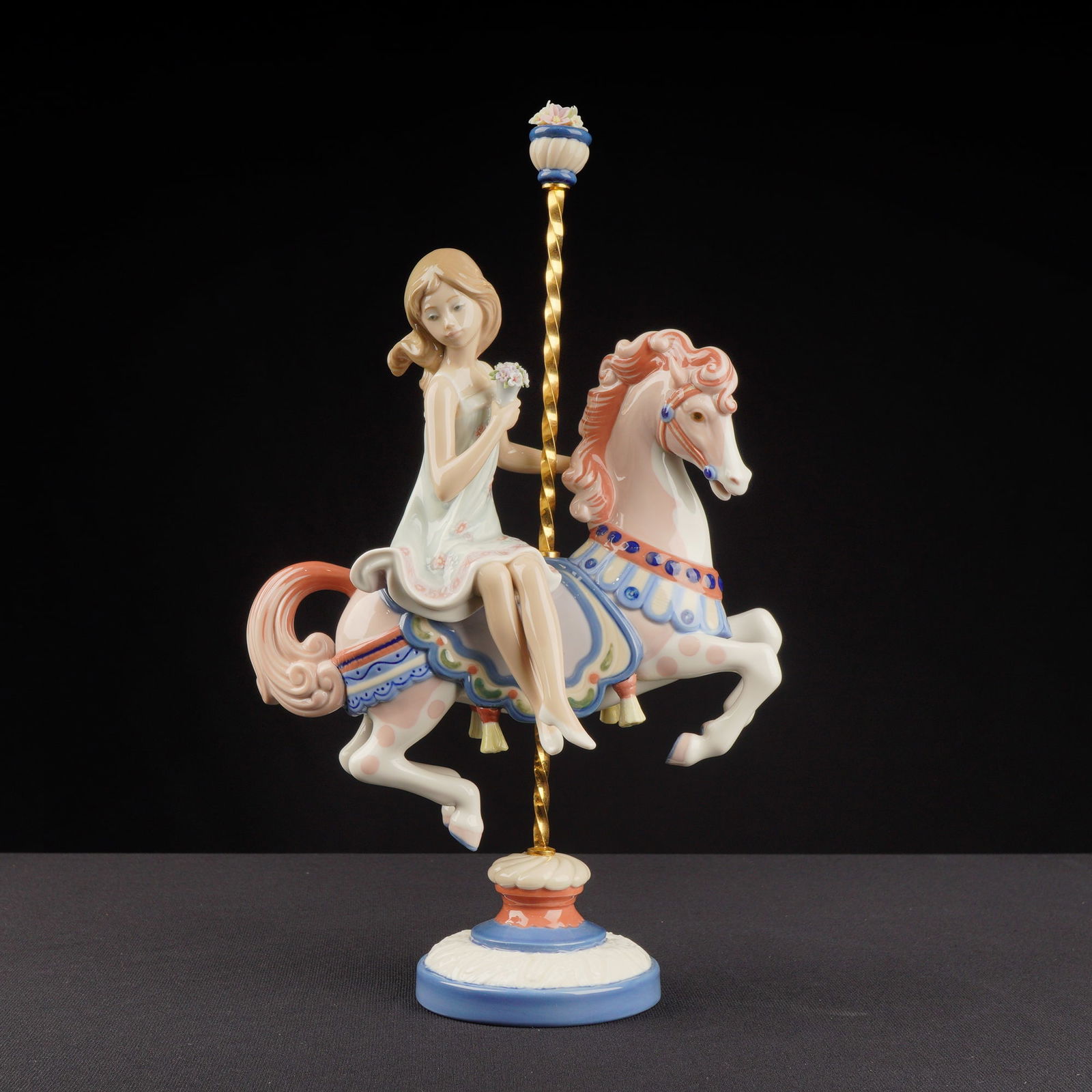 Lladro Porcelain Sculpture Girl on Carousel Horse 01001469: The glazed porcelain figurine depicts a young woman in a pale dress seated upon a decorative carousel horse. The sculpture is mounted on a central, twisted gold-toned metal pole which rests on a stepp