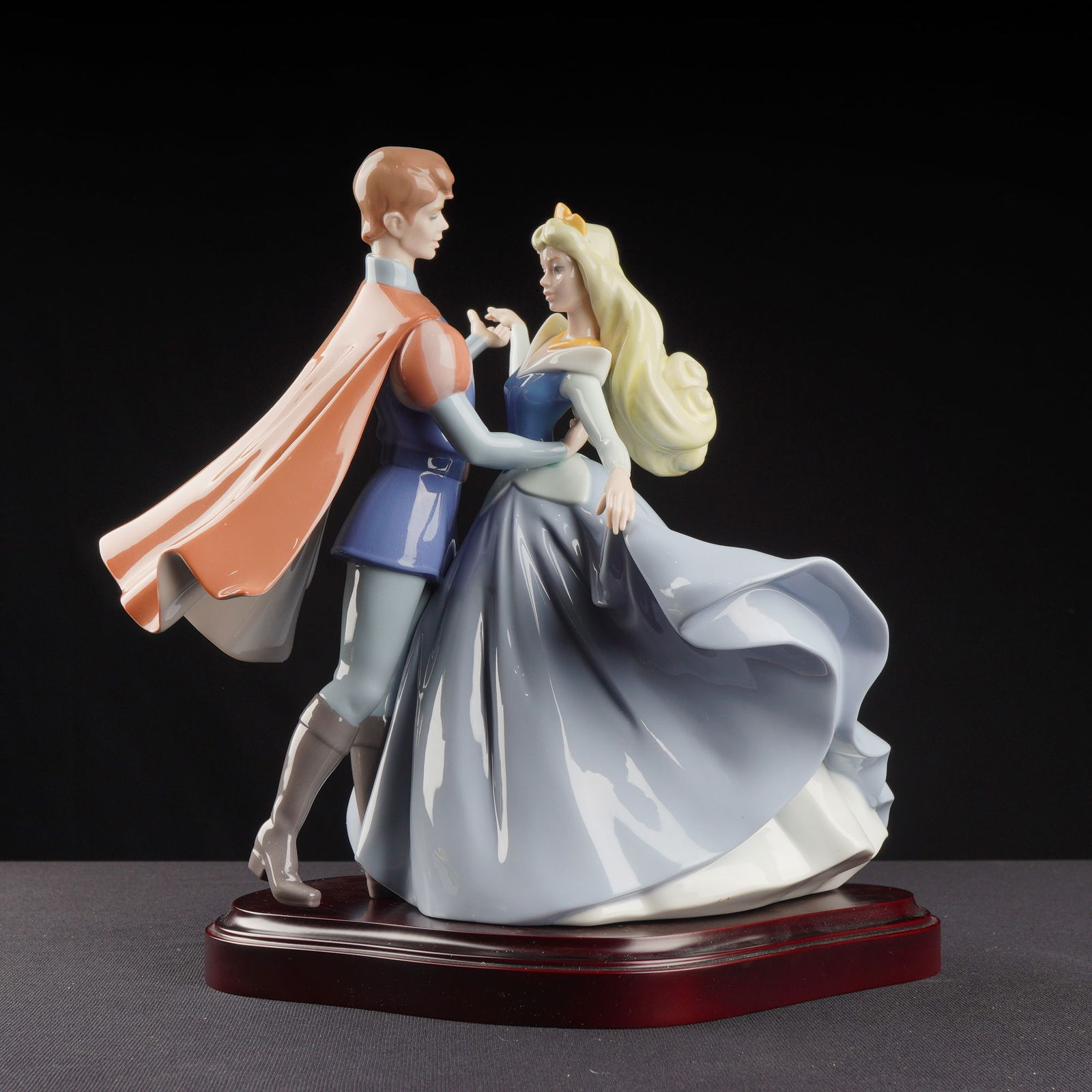 Lladro Porcelain Sculpture, Sleeping Beauty's Dance 01007560 with Base: This limited-edition piece number 552 of 1000, depicts the Disney characters Princess Aurora and Prince Phillip engaged in a dance, mounted on a polished, reddish-brown wooden base. The figurine is cr