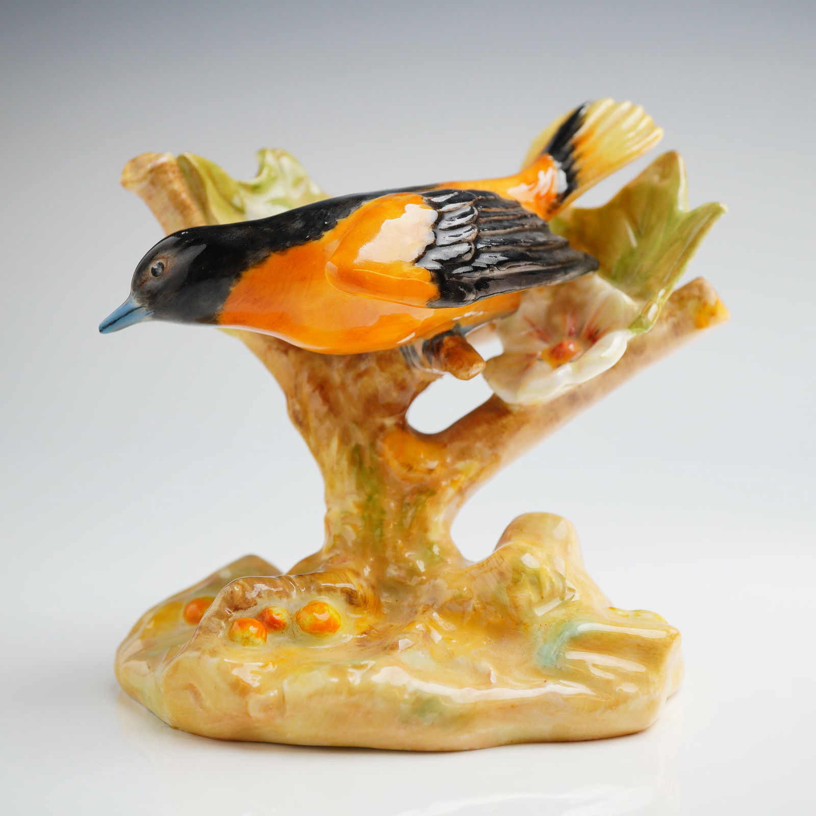 Royal Doulton Bone China Figurine Baltimore Oriole HN 2542: A single glazed bone china figurine by Royal Doulton, depicting a Baltimore Oriole perched on a stylized tree branch. The bird features black coloring on the head and wings, contrasting with vibrant o